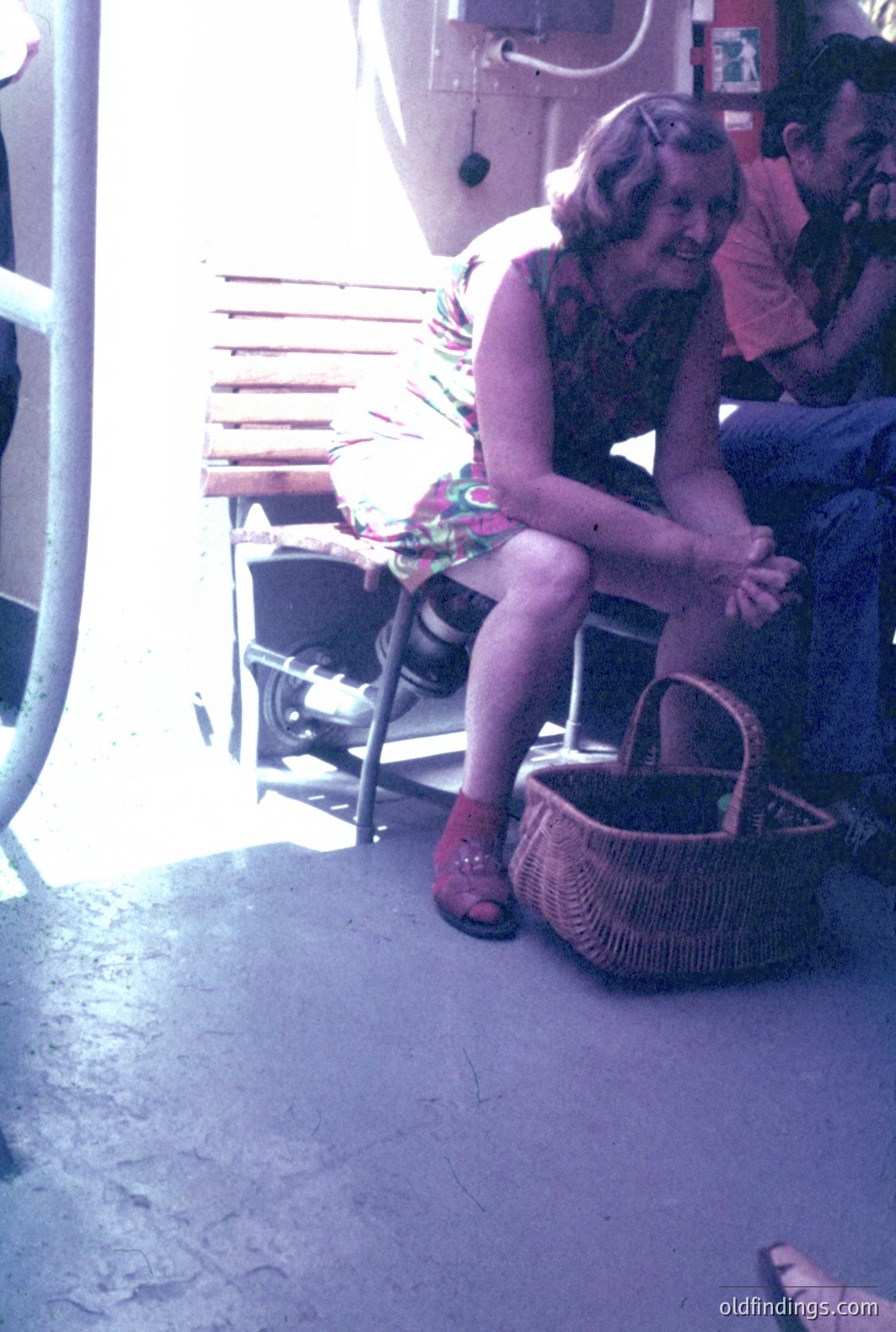 A woman with shoulder-length hair and a floral print dress sits beside a woven basket. A man in jeans and an orange shirt is partially visible. Appears to be a boat deck, likely a ferry or cruise ship, based on industrial piping. Color photo, likely 1970s. Shows everyday leisure activity.