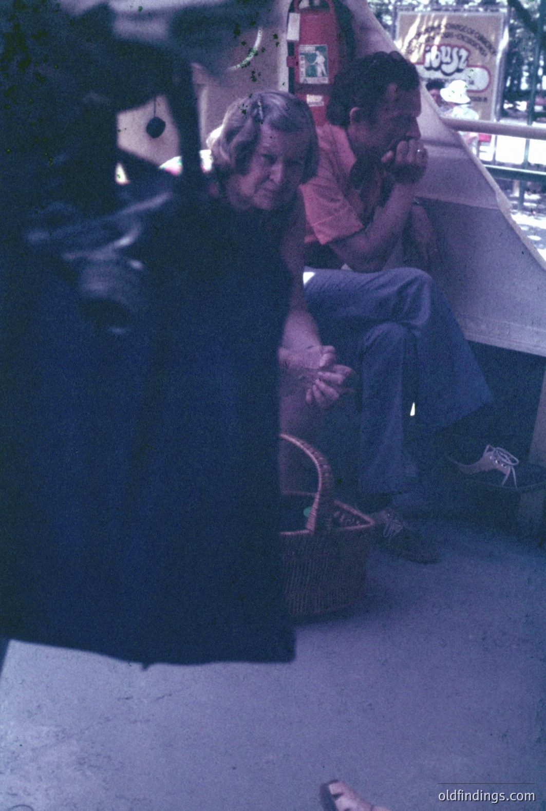 A young boy, circa 1970s, sits in jeans and a red t-shirt on a concrete ledge. A woman with long hair wears a dark dress & sandals, appearing to comfort or console him. The scene's backdrop features blurred, colorful signage. Possible outdoor market setting. Evokes a candid, intimate moment.
