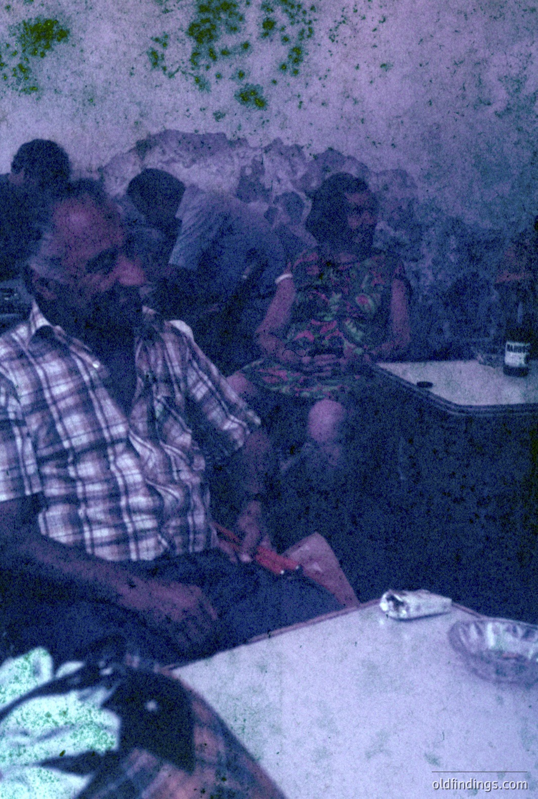 A candid shot captures two people seated at a table with a patterned tablecloth, likely in a café. The man, with a full beard, wears a plaid shirt, while the woman has on a floral dress. A cigarette rests on the table, suggesting a casual atmosphere. Appears to be a snapshot from the 1970s.