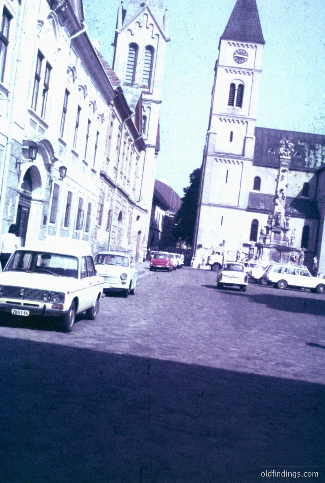A wide, cobblestone street leads toward a towering church with a pointed steeple and ornate facade. Vintage cars, including a Fiat, line the road. Building architecture shows Baroque influences. Likely a European town, likely 1970s. A snapshot of everyday life.