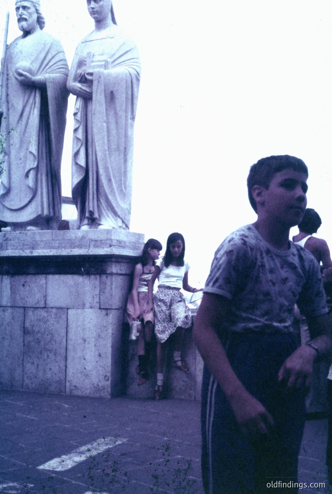 A candid street scene showing a young boy in casual clothing and two girls standing near a monumental statue, likely depicting historical figures. The image exhibits a faded, possibly color-shifted aesthetic suggesting a 1970s or 1980s origin. Appears to be a public square or plaza.