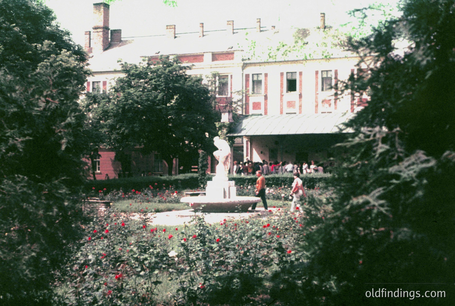A formal garden with a central fountain featuring a classical statue, framed by dense foliage. A large, stately building with multiple arched windows and a distinctive roofline dominates the background. People are visible near the building's entrance. Appears to be a public park or estate. Likely 1970s or earlier.