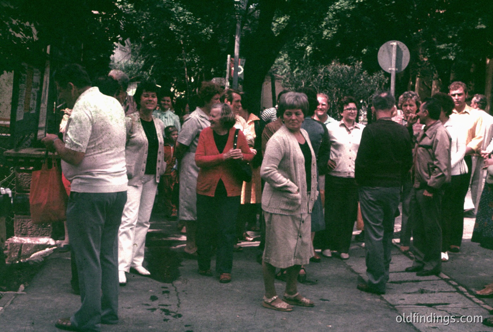 A group of people gathers outdoors on what appears to be a street or promenade, likely in a European coastal setting. Fashion suggests the 1970s. Visible architecture includes a streetlight and signs. Several women wear distinctive hairstyles & clothing.