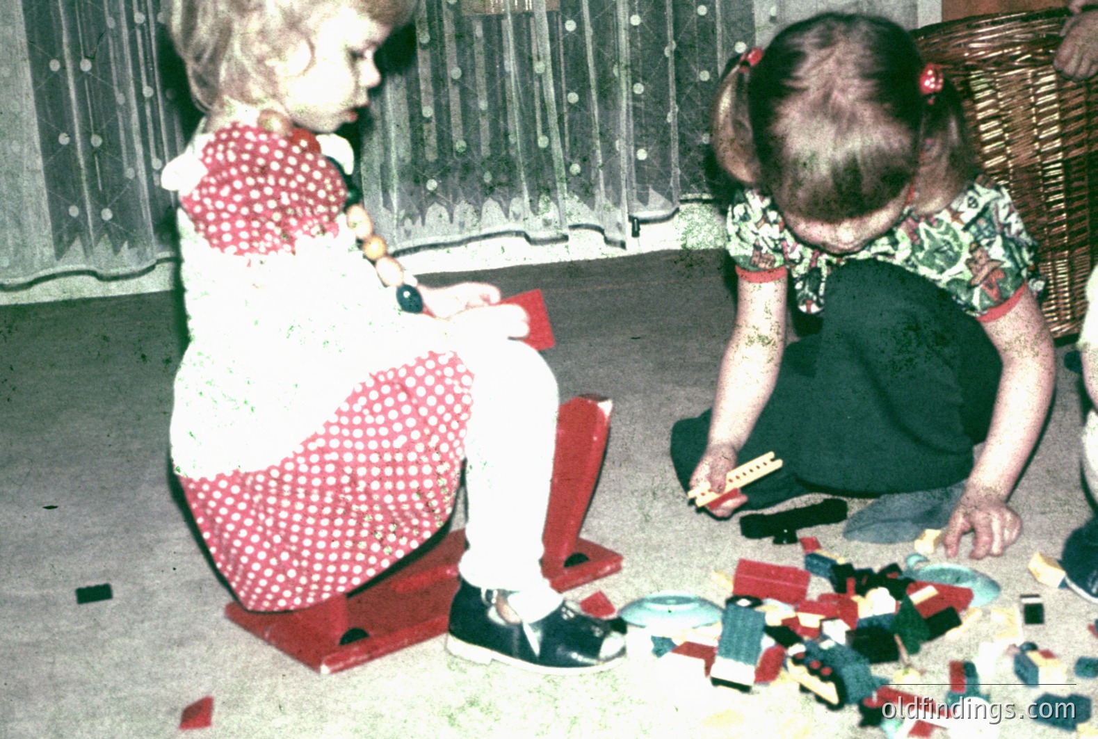 Two young girls play with toys in a warmly lit, domestic setting. One sits atop a red plastic toy, wearing a red-and-white patterned dress and a beaded necklace. The other is engrossed in building with colorful plastic blocks. A wicker basket sits nearby. Likely a family snapshot, mid-1970s.