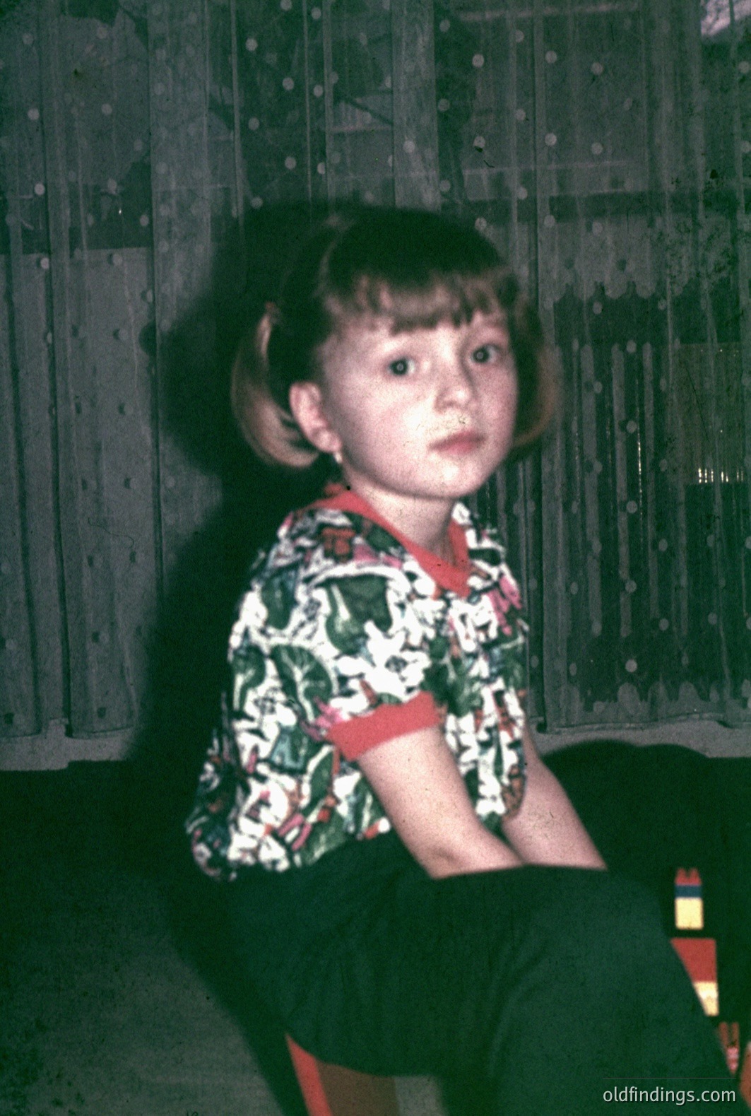 A young girl with a bowl cut sits indoors, gazing directly at the camera. She wears a vibrant, patterned shirt with red trim and dark-colored pants with unique designs. The backdrop features patterned drapes. Likely a casual family snapshot, circa 1970s.