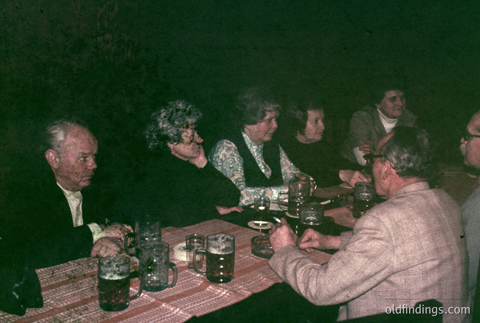 A group gathers around a table, likely enjoying a meal or drinks. Visible are several older adults; men in suits and sweaters, women in patterned tops and dark jackets. Mugs and glassware suggest a casual gathering, potentially in a dimly lit, rustic setting. Likely 1970s-80s era.