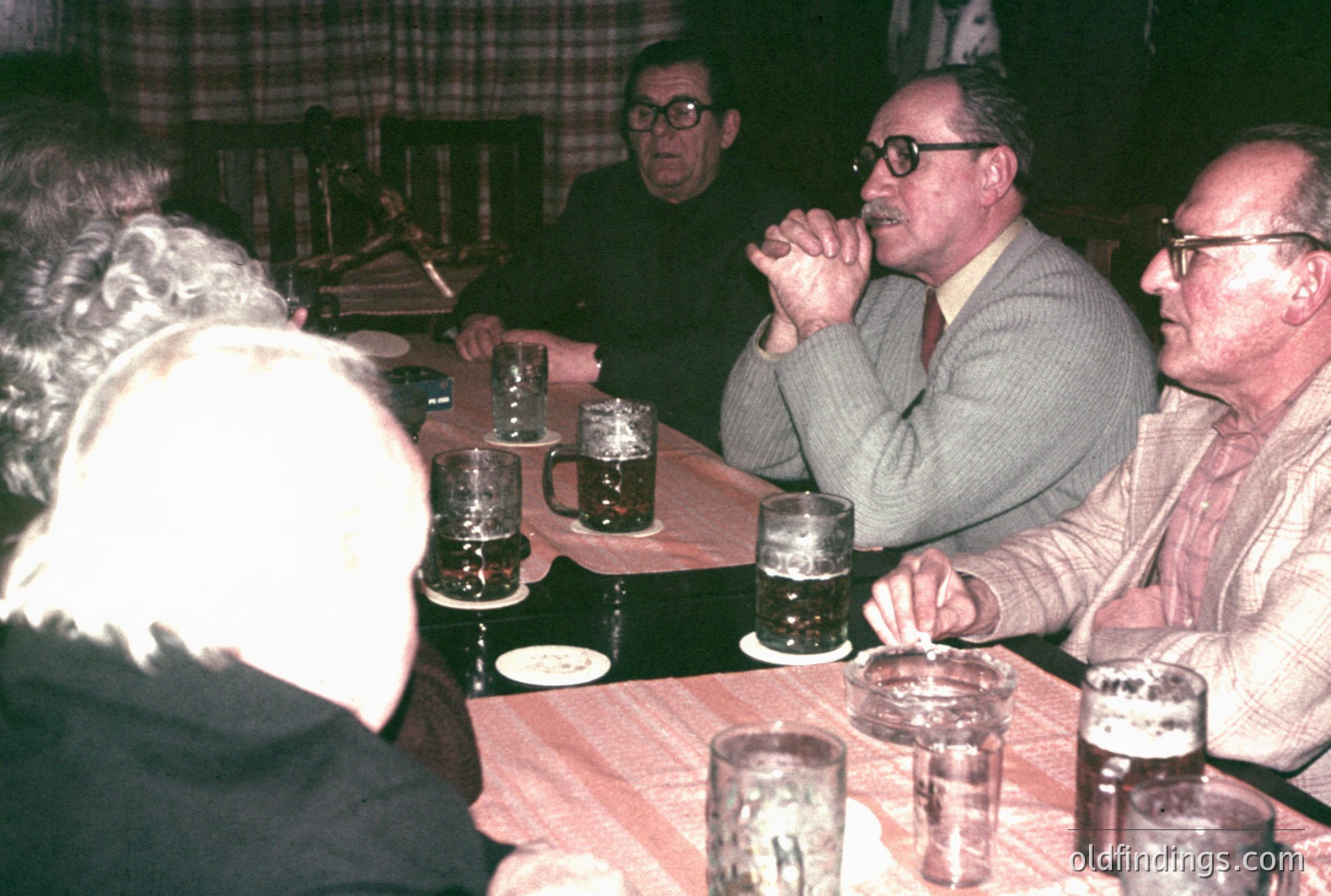 Four men seated around a table covered with a red checkered tablecloth. Several beer steins and ashtrays are visible. The men are wearing 1970s-era attire with glasses and mustaches. Casual, intimate gathering. Likely a social club or tavern setting. Warm, nostalgic color palette.