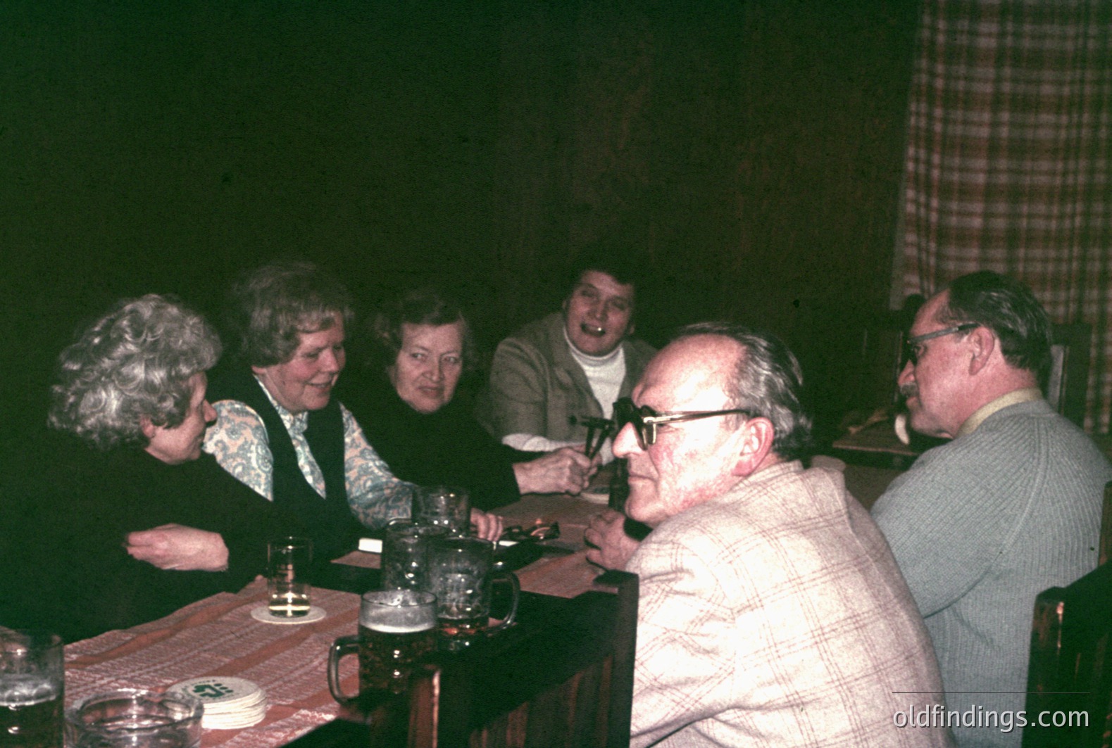 A group of five adults gathered around a wooden table in a dimly lit room. Floral and patterned attire suggest a casual, homey setting. The room's dark tones and dated decor hint at a 1970s timeframe. Likely a candid snapshot of a family or friends' gathering.