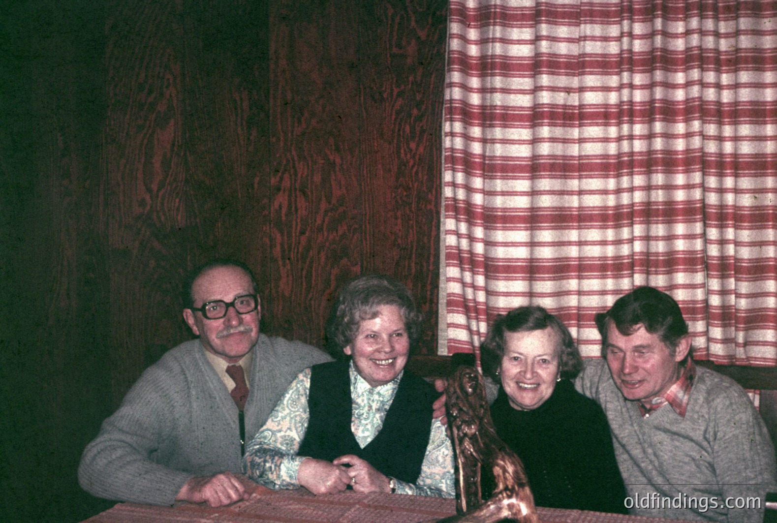 Four individuals seated at a table covered with red fabric, posed for a portrait. The décor includes wood-paneled walls and a red and white checkered curtain. Likely a family gathering or celebratory event, c. 1970s. A small statue sits on the table.