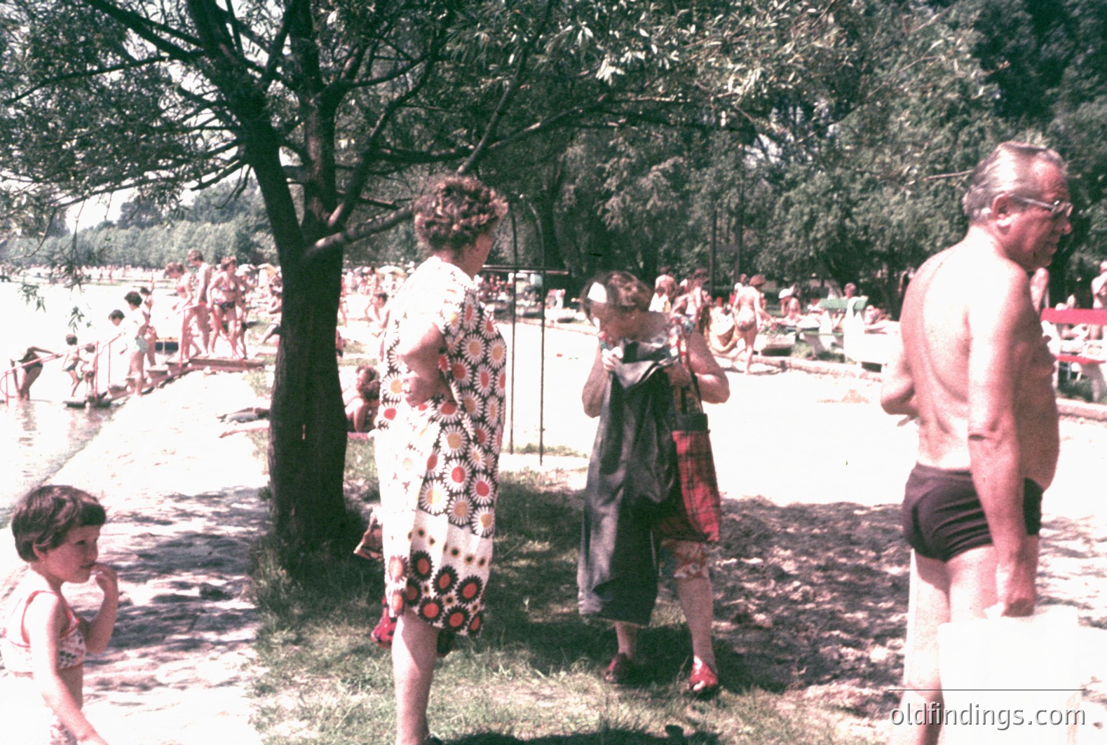 A lakeside scene with families enjoying a summer day. A young girl stands near a woman in a 1960s-70s patterned dress & hat. A man in swimming trunks stands nearby. Background shows swimmers and lush foliage. Likely Eastern Europe. Vintage color photography.
