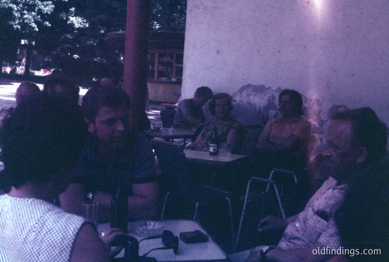 Group of men and women seated at tables outdoors; likely a cafe or restaurant patio. The scene shows casual clothing, hairstyles typical of the 1970s. Possible seaside location with faint glimpse of water in background. Color palette is muted, indicative of early color photography.