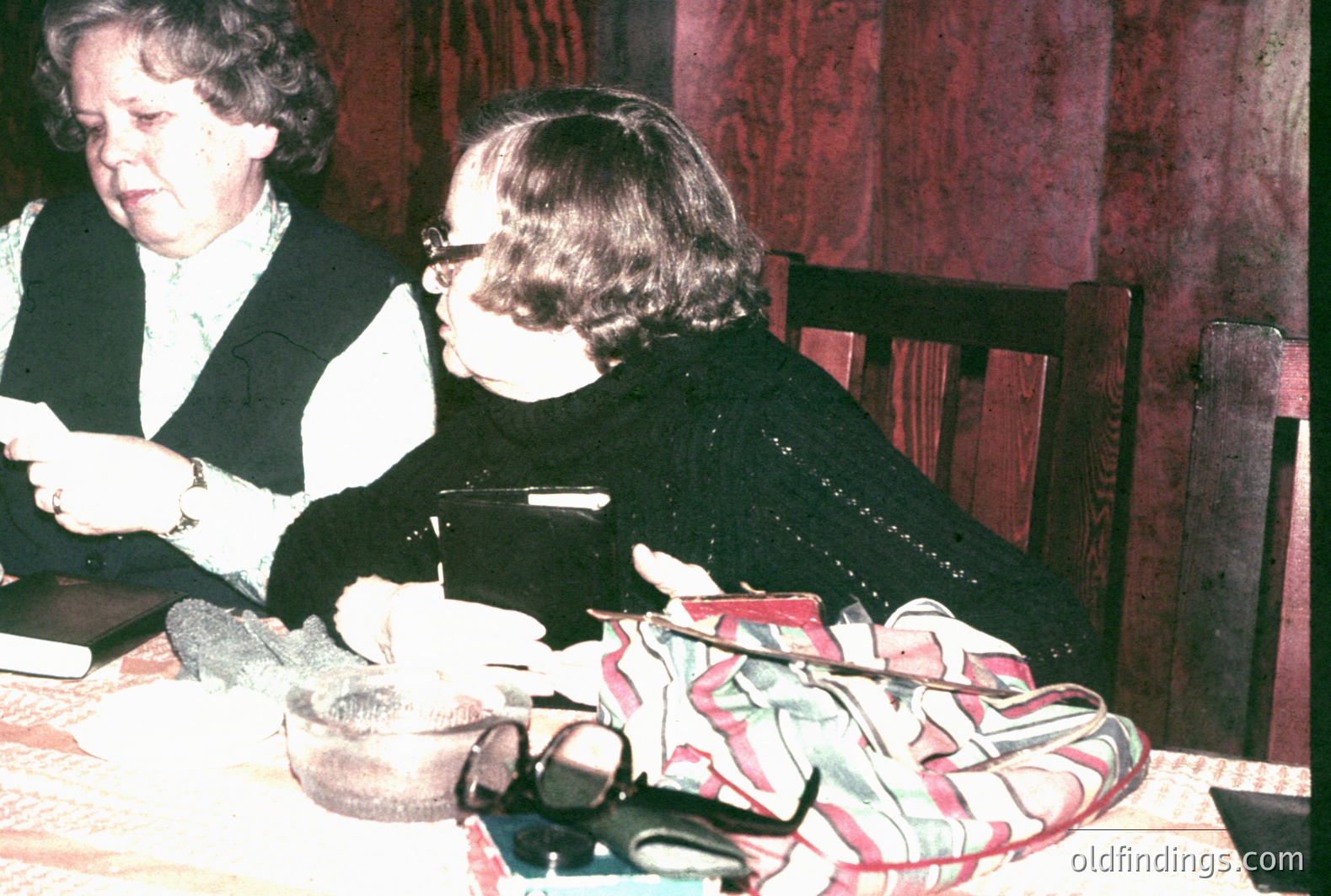 Two women are seated at a table, appearing to be reviewing documents. One wears a crisp white shirt and a vest. The other is in a dark sweater. A patterned bag sits on the chair beside her. Likely a candid moment. Appears to be from the 1980s due to color palette and clothing styles.