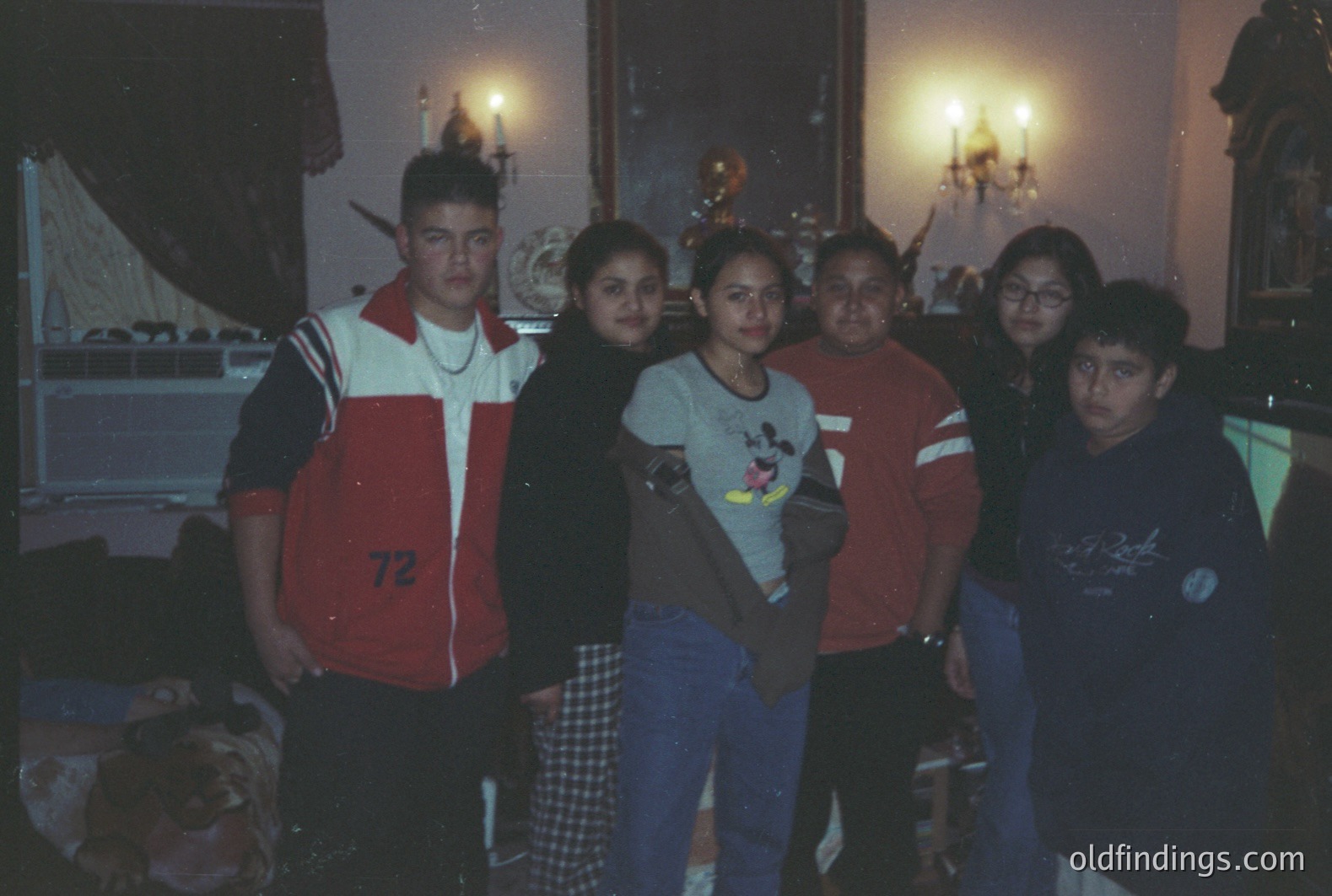 Group portrait of five young people in a richly decorated room. Features include a patterned rug, ornate wallpaper, and lit candelabras. Fashion suggests late 1990s/early 2000s – track jacket, jeans, graphic tees. Casual, snapshot aesthetic. Likely a family gathering or event.