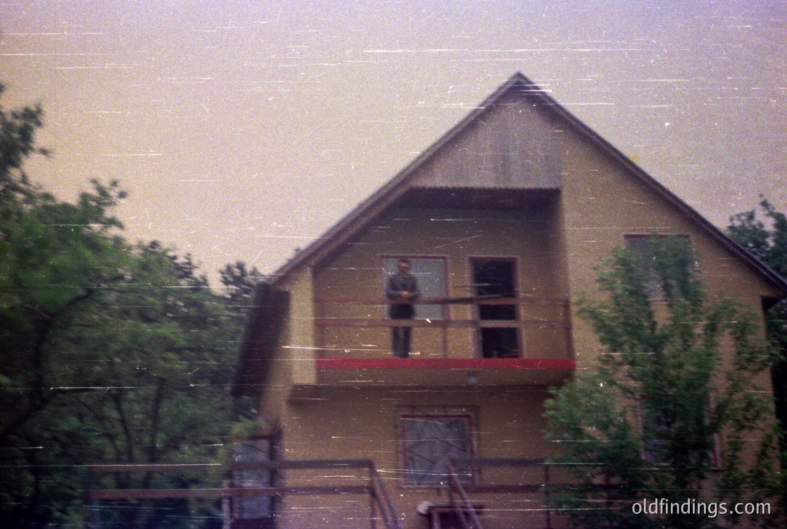 A two-story building, seemingly residential, features a prominent balcony with a single figure visible. The architecture suggests a mid-century aesthetic. The image exhibits film grain and a slightly faded color palette, implying a vintage photograph. Lush foliage frames the building. Appears to be taken in Bulgaria.