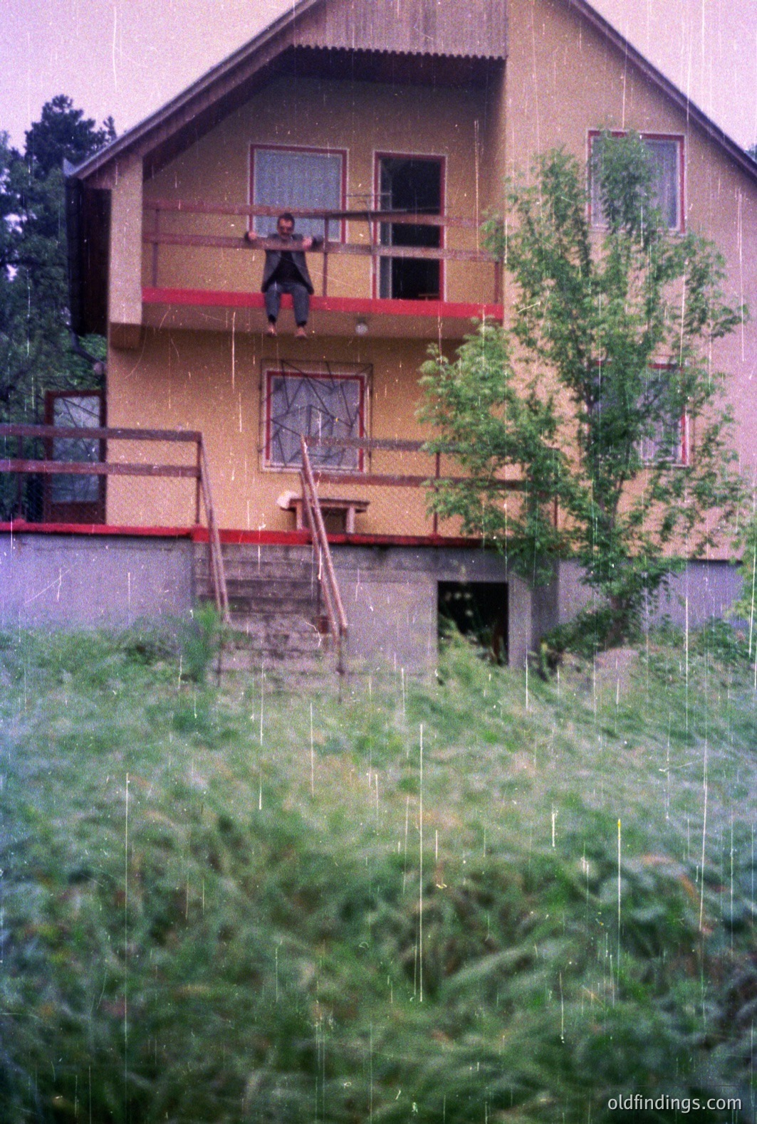 A two-story building with a balcony featuring a person seated and facing away. Visible architectural details include a red-trimmed balcony and staircase. Dense foliage obscures the ground level, suggesting a rural or semi-rural location. Rain streaks the image. Likely 1970s or 80s era.