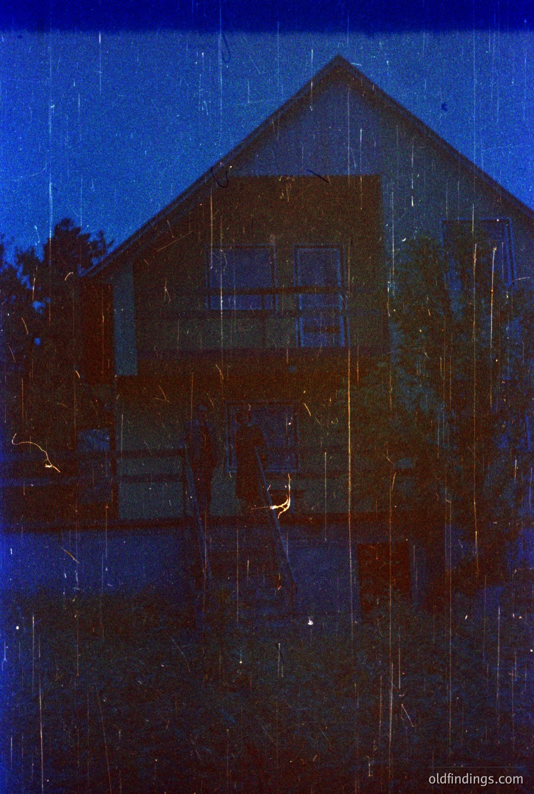 Striking blue tint photography depicting a two-story, wood-framed house with a prominent staircase. Visible details suggest a mid-century modern architectural style. Grainy texture and apparent light leaks indicate potential age/vintage film stock. Atmospheric and evocative, potentially useful for design.
