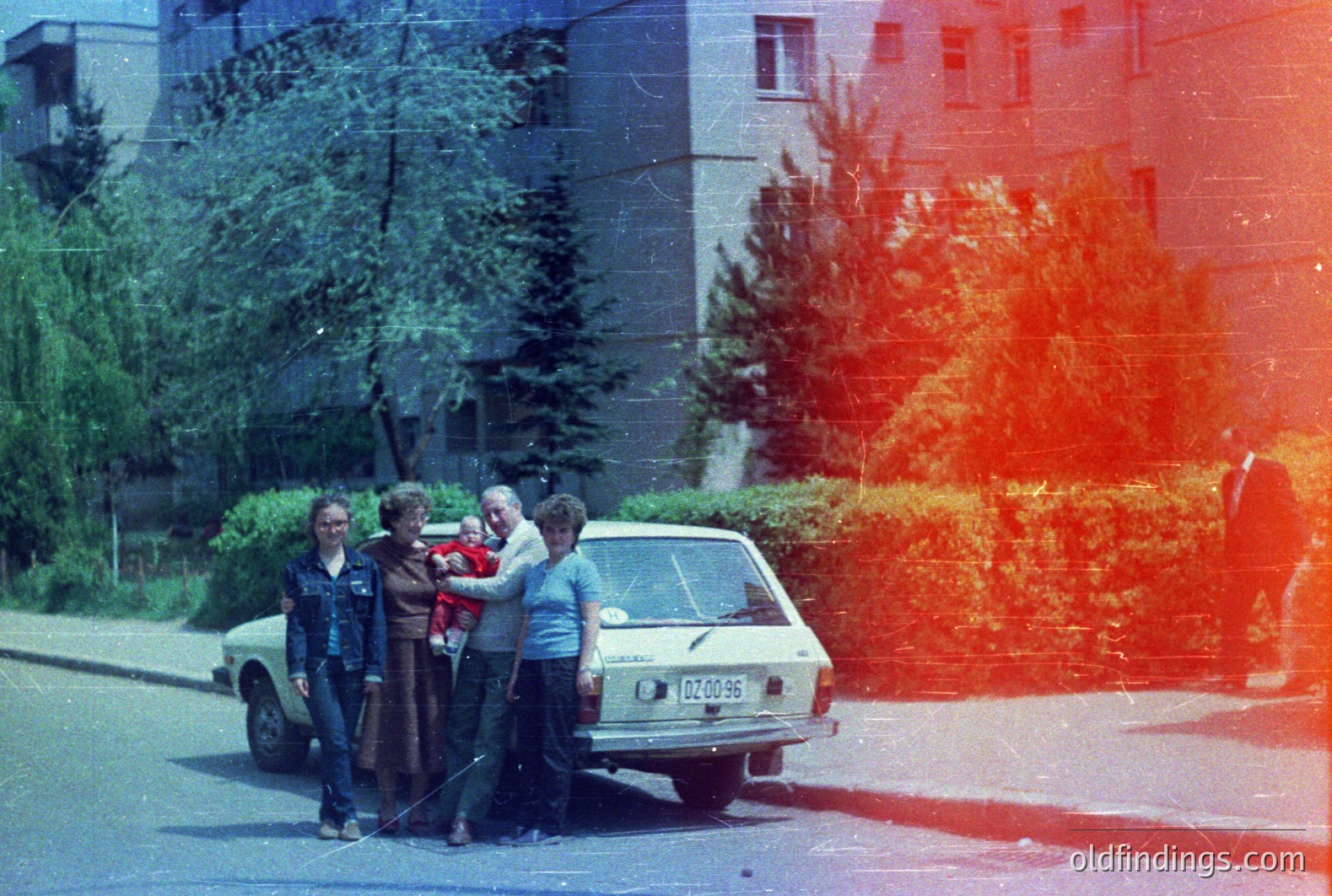 A family poses beside a light-colored sedan, likely a Dacia 1300, near an apartment block. Three individuals, a woman holding a baby, a man, and a teenager stand on a paved road lined with greenery. The architectural style suggests Eastern Europe, potentially Bulgaria. 1970s aesthetic, likely color slide film. Automotive and family portrait reference.