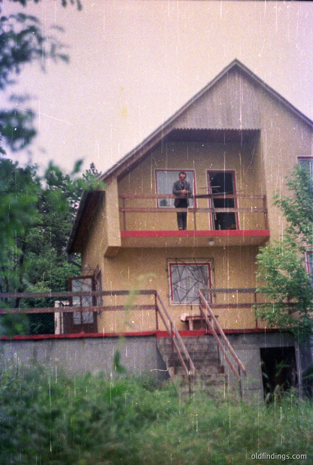 A two-story building with a prominent balcony features a man standing nearby. The structure showcases a mix of architectural styles, with a steeply pitched roof and red-accented railings. Appears to be a resort setting. Likely 1970s-1980s construction. Notable photographic style.