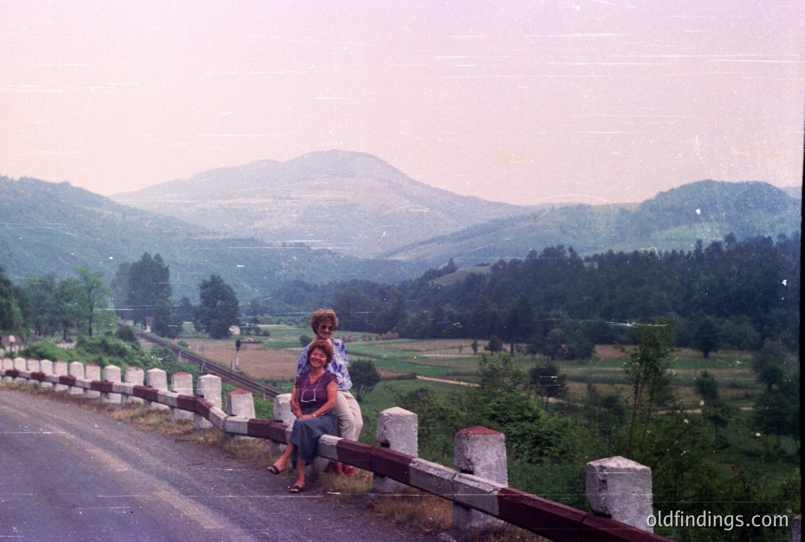 Two women seated on a roadside barrier overlooking a valley of lush, forested hills. View is dominated by a distant, hazy mountain peak. Appears to be a rural, possibly Balkan region, possibly 1970s film photography aesthetic. A classic travel snapshot.