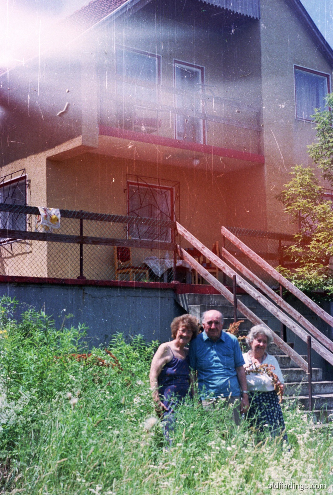 A family stands on a grassy slope in front of a multi-story apartment building. The architecture suggests a Central European or Alpine style. The building features a balcony with red trim and a steeply pitched roof. Likely taken in the 1970s or 1980s, indicated by the film aesthetic & clothing styles. A vintage, slightly faded color print.
