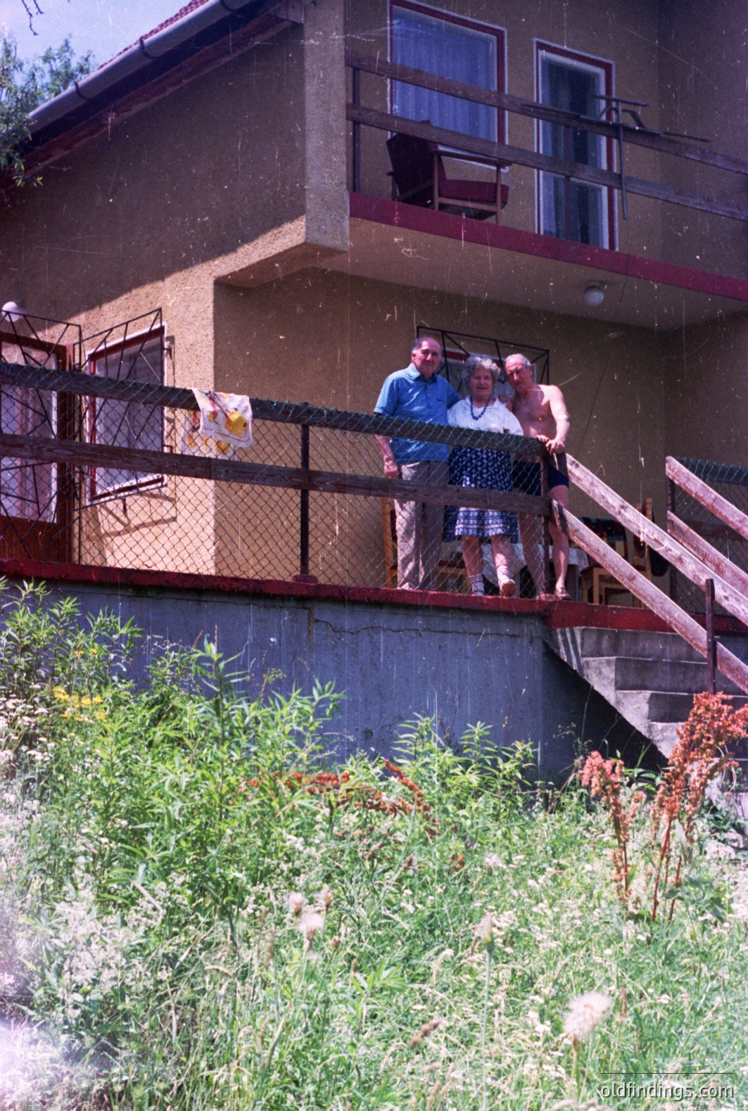 Three people stand on a balcony of a concrete building, likely a seaside resort. The building features 1970s-era architecture with a projecting balcony. Viewers appear to be on holiday. Lush foliage fills the foreground. A glimpse of the Black Sea is visible between buildings.