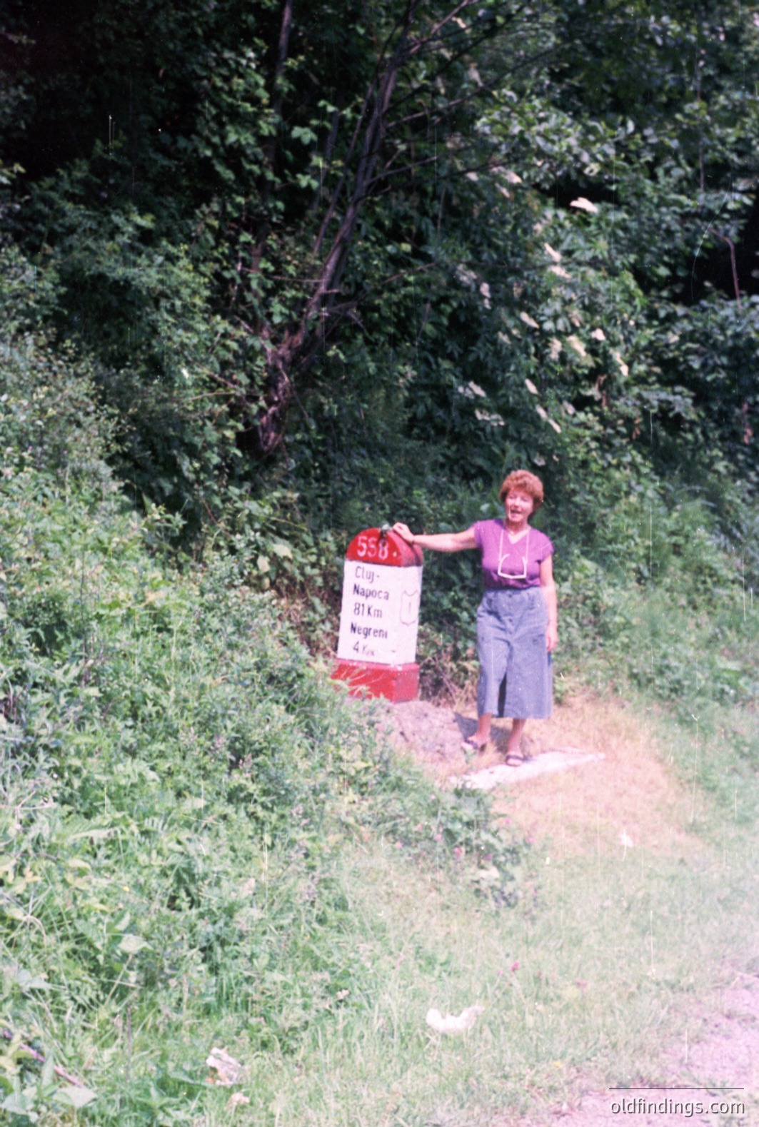 A woman stands next to a signpost in a rural setting. The sign, likely indicating road distances, is in a non-Latin script (possibly Cyrillic). Vegetation heavily populates the background and foreground. The style suggests a casual snapshot, likely from the 1970s or 1980s, potentially in Eastern Europe (Bulgaria?).