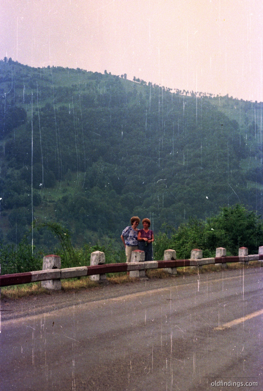 Two figures stand near a roadside barrier overlooking a heavily forested hillside under a rainfall. The scene suggests a mountain pass or scenic overlook. Likely a travel snapshot, possibly 1970s or 1980s, judging by the film quality and clothing styles. The image evokes a sense of adventure and exploration.