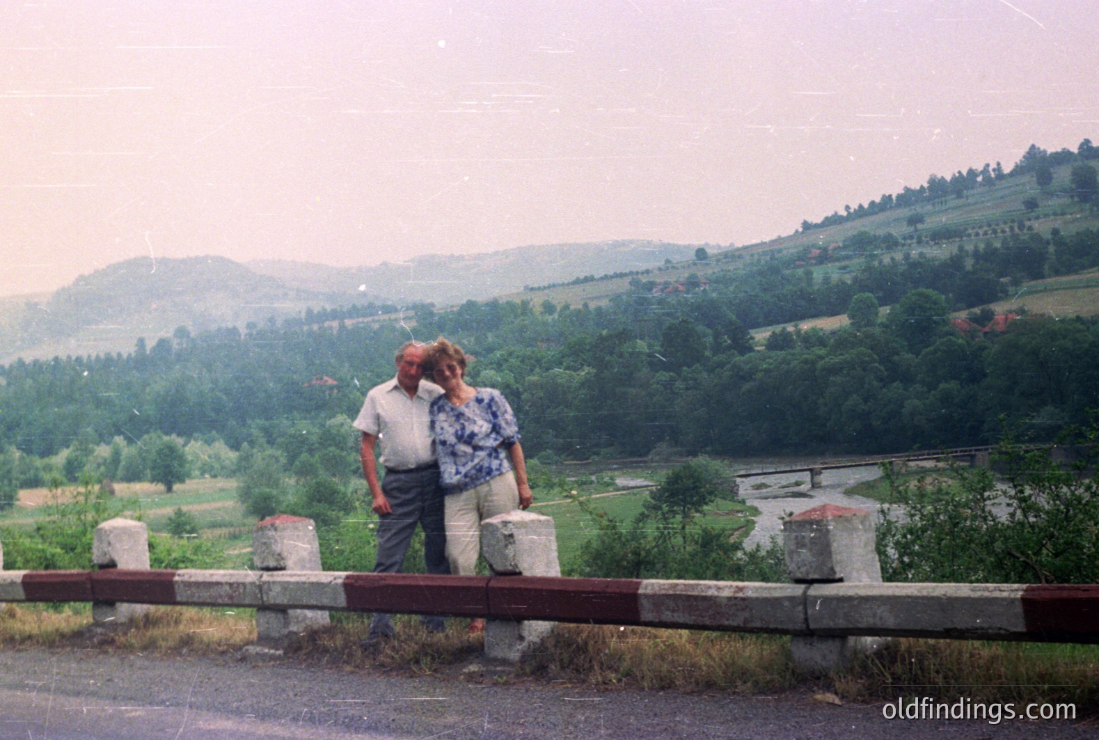 An older couple stands behind a road safety barrier, overlooking a forested valley with a river winding through it. Man wears a short-sleeved shirt; woman in a light-colored top. Likely taken in the 1970s or 80s given the photographic style & clothing. Rural landscape, potentially Eastern Europe. Commercial value: travel, family, nostalgia.