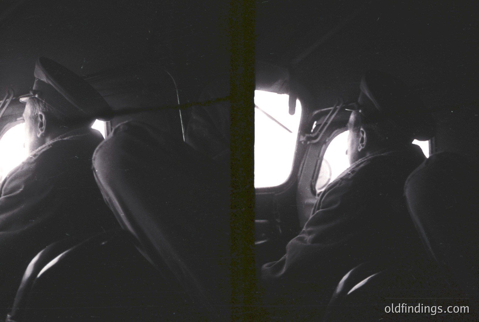 A man in a dark coat and hat sits facing away from the camera, viewed from behind. The scene is brightly lit with light emanating through a window, creating a strong contrast. Grainy texture suggests film photography. Likely a candid street portrait, possibly 1960s-1970s.