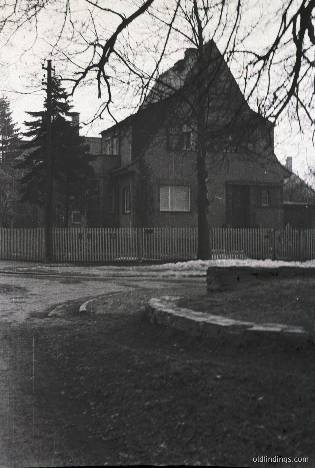 A two-story residence with a steeply pitched roof and symmetrical facade. Features include small, rectangular windows and a split-rail fence along the front property line. Possibly a suburban or rural setting, indicated by surrounding trees and a winding road. Likely taken in the mid-20th century.