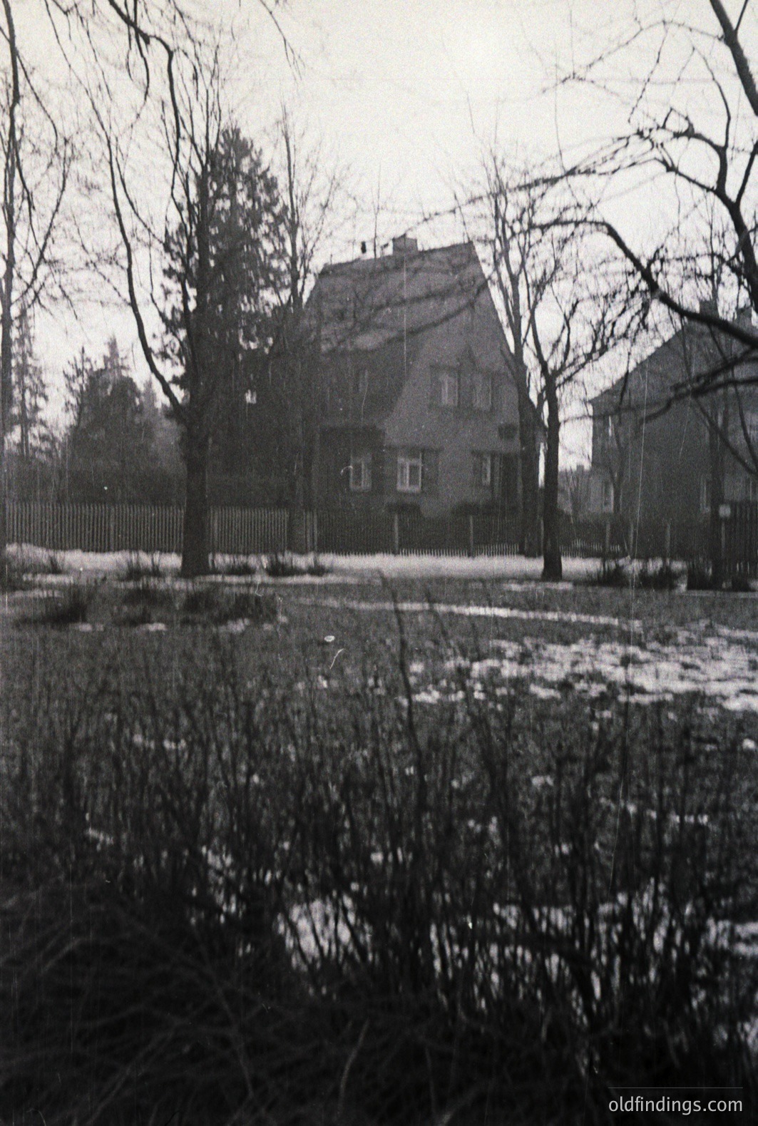An austere, two-story home with a steeply pitched roof and prominent dormer window dominates the frame. Sparse ground cover and bare trees suggest a late autumn or early winter scene. Likely a residential neighborhood, early-mid 20th century. Architectural style is simple, functional.