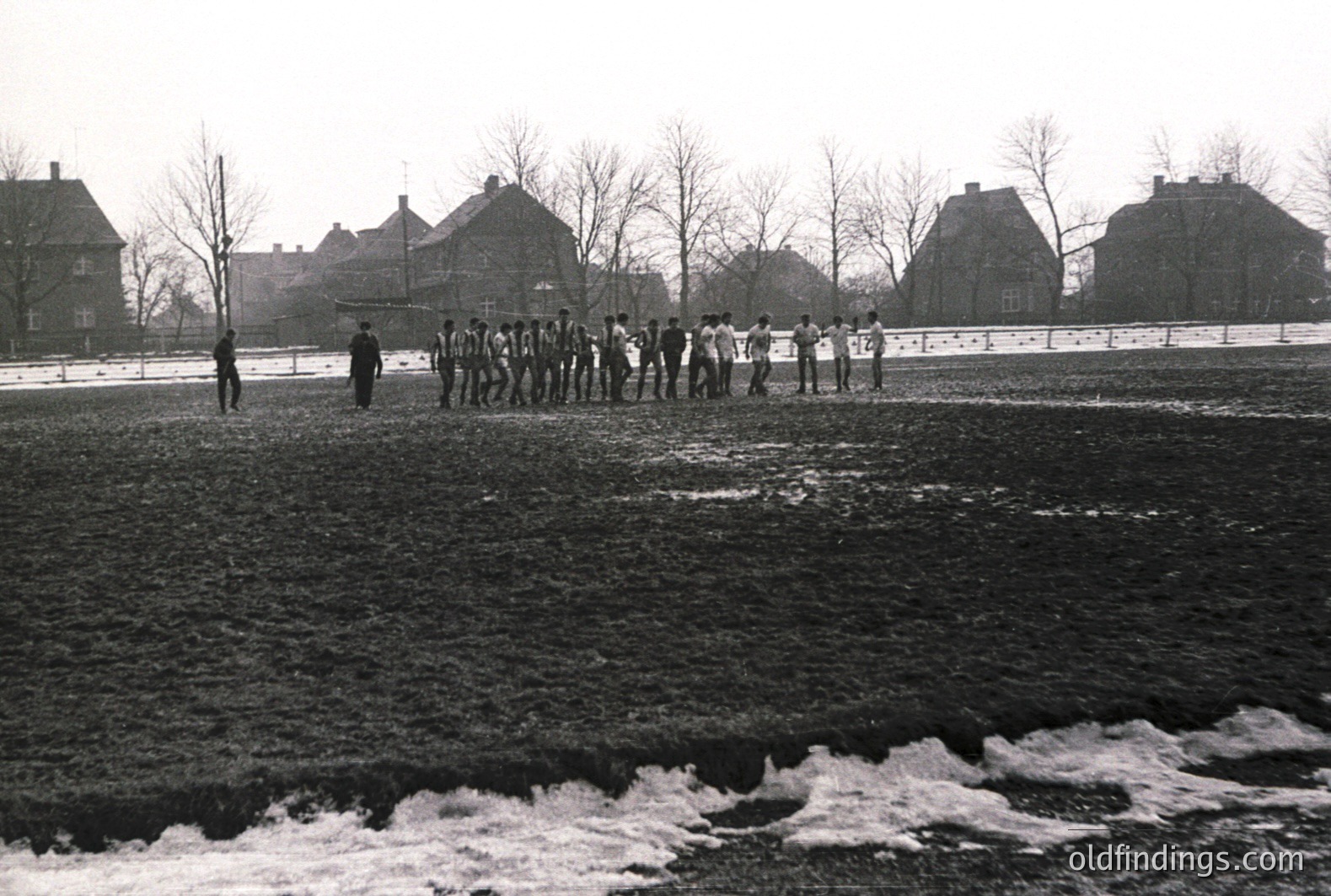 A group of young men, possibly a sports team, stand in a line on a muddy field with a row of brick buildings in the background. The scene suggests a chilly, overcast day; patches of ice are visible. Likely a European location, mid-20th century. Appears staged, possibly for a team photo.