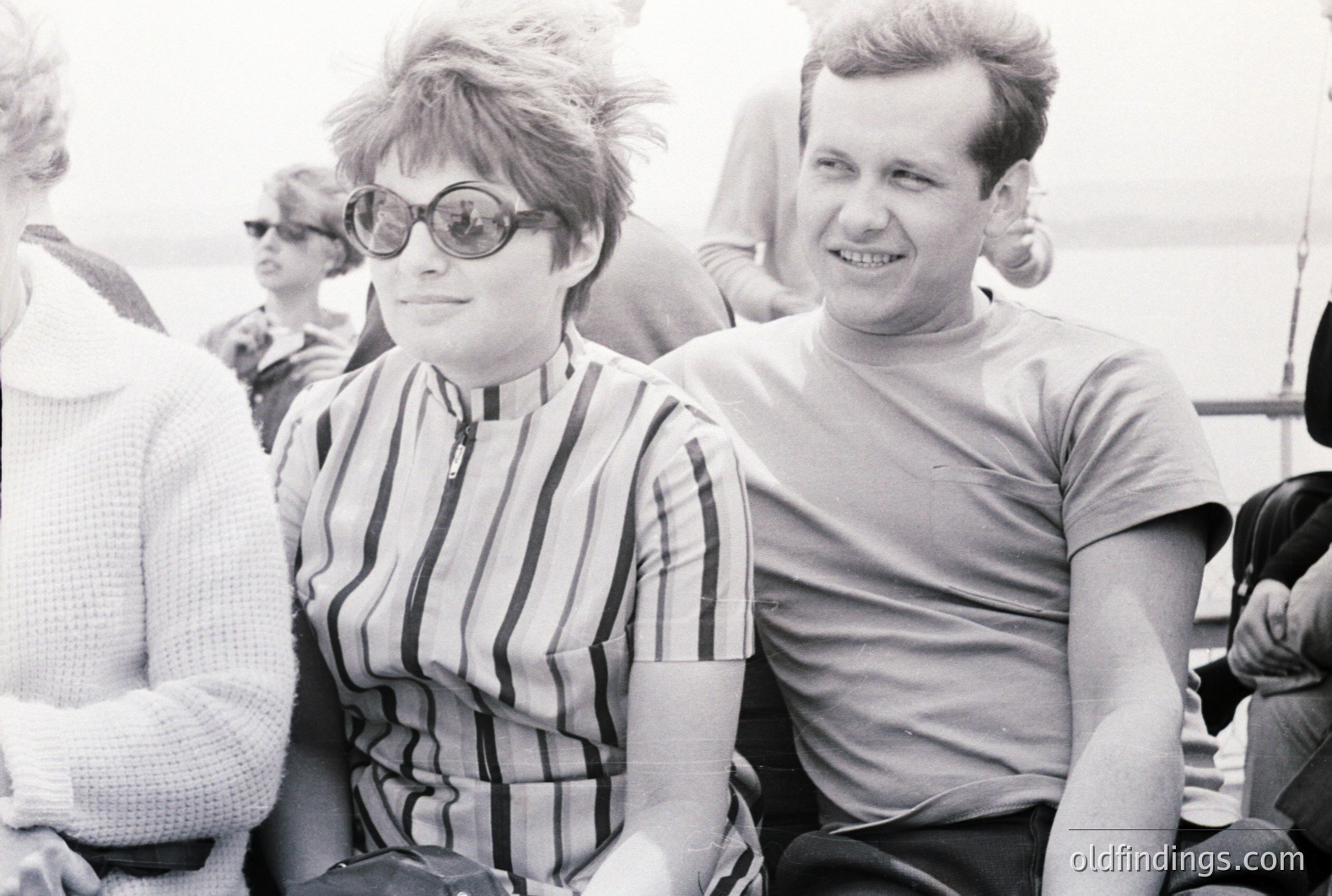 A black and white portrait captures a young couple, seemingly on a boat. The woman wears a striped high-neck top & distinctive round sunglasses, and has a stylish bob. The man smiles; both appear relaxed and casually dressed. Likely 1960s style. Background shows blurred figures and water.