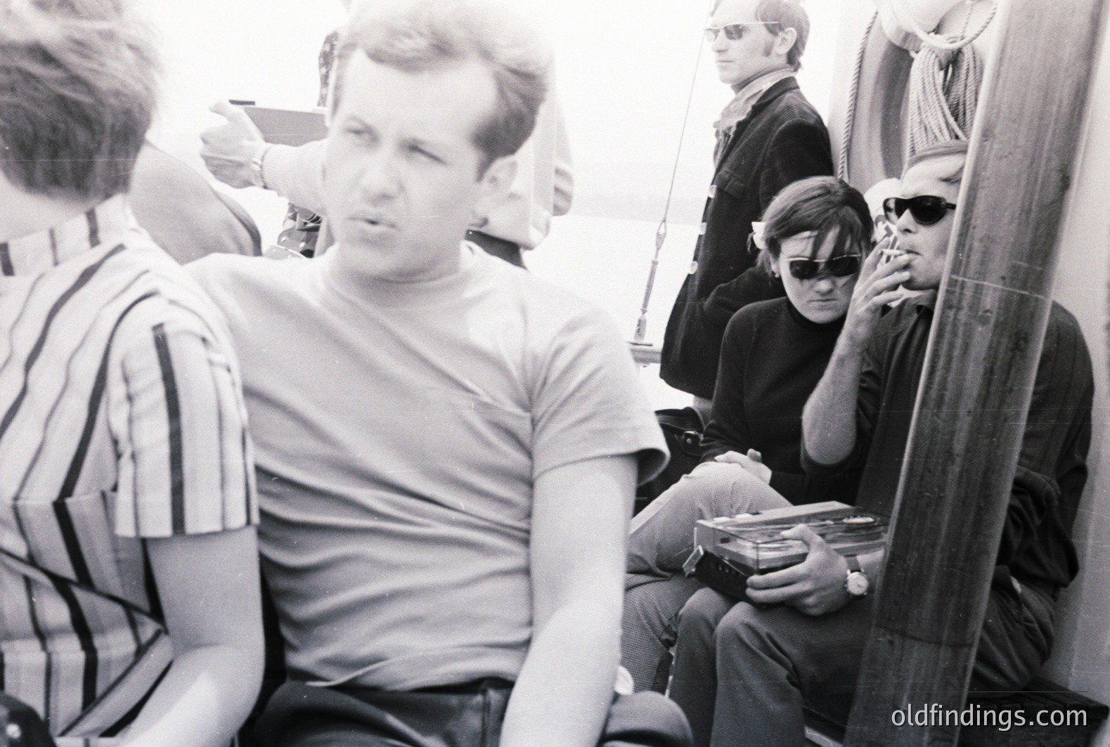 Three men aboard a sailboat, appearing relaxed and engaged in smoking. The central man wears a simple t-shirt, while others sport dark sunglasses and tailored clothing indicative of the 1960s. Visible nautical ropes and rigging establish the setting. Likely promotional material for leisure or travel.