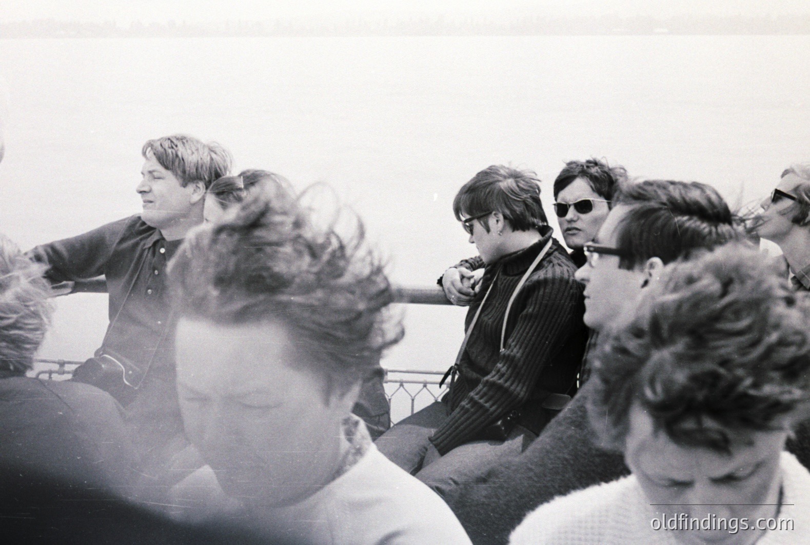 Group of young men, likely tourists, are seated on a boat deck, gazing towards the horizon. Dark, casual clothing and hairstyles suggest the late 1960s or early 1970s. Black and white photography, grainy texture. A sense of leisure and travel.