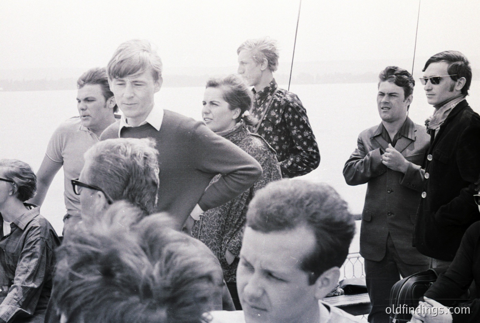 A group of formally dressed people are gathered on a boat deck overlooking a body of water. The scene, likely captured in the 1960s, shows men in suits and sweaters and a woman in patterned fabric. A distinctive nautical railing is visible. Possible leisure or business travel.