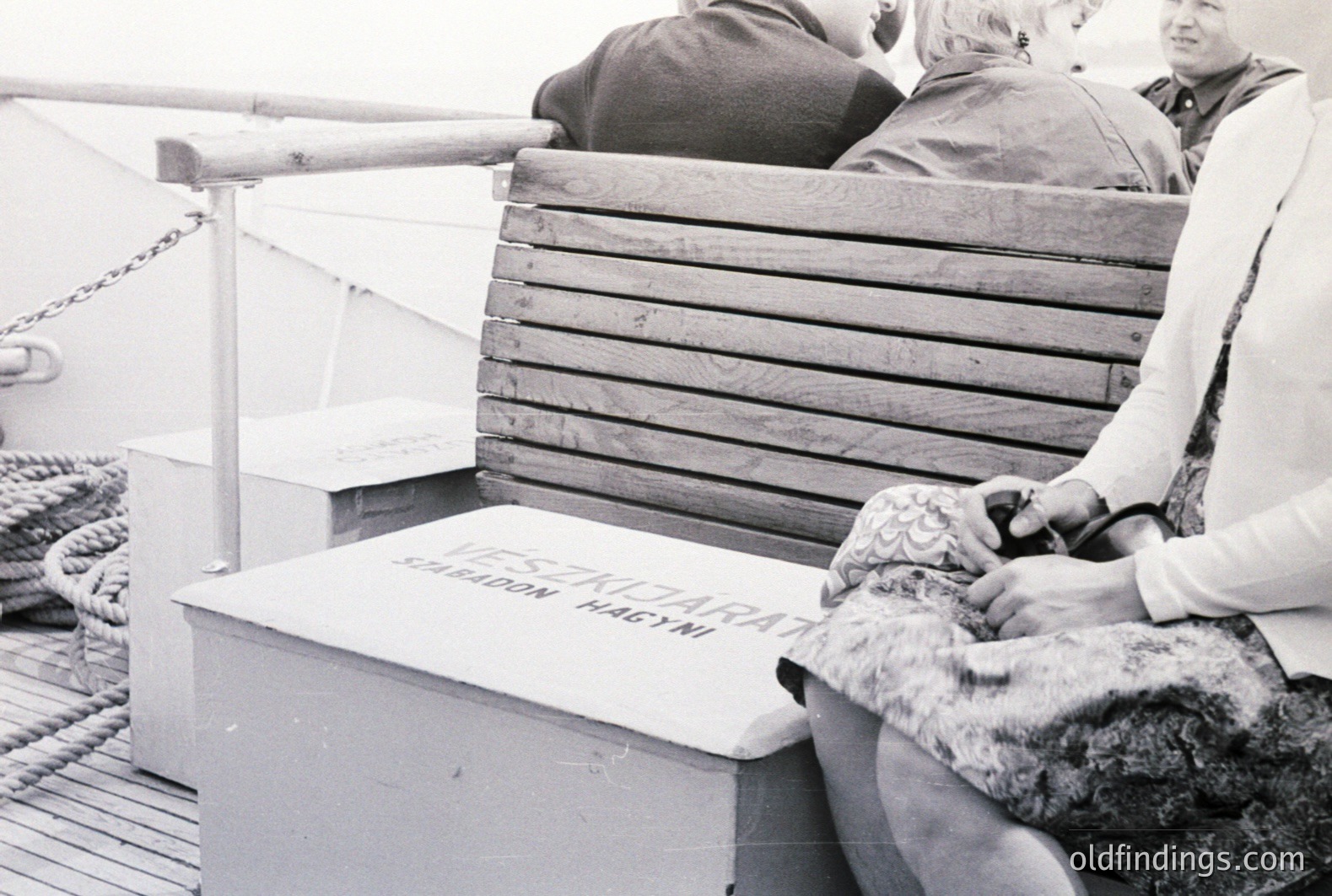 Monochrome photo depicts three figures seated on ship deck. Two women in patterned dresses and a man wearing a jacket. A weathered wooden bench with Cyrillic lettering is prominent. Likely 1960s-70s era. May reflect travel, leisure, or maritime history.