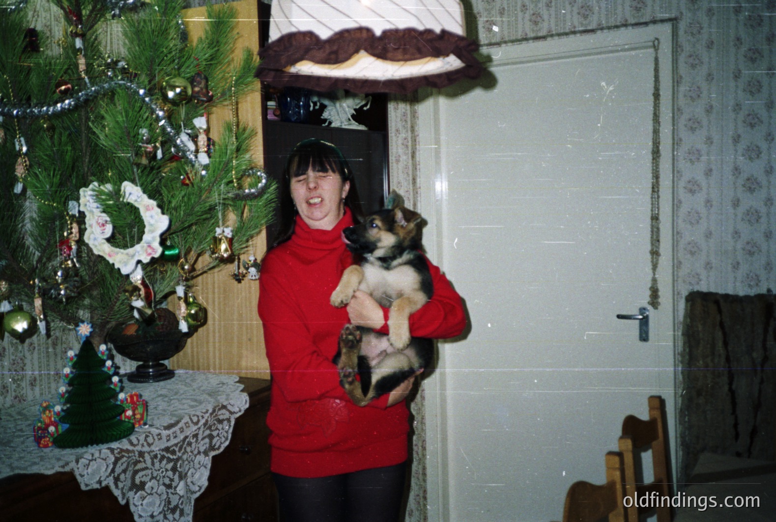 A young woman cradles a German Shepherd puppy in a warmly-lit interior. A decorated Christmas tree stands behind her. The image exhibits characteristics of 1970s or 80s photography—color saturation and film grain. Likely a family snapshot.