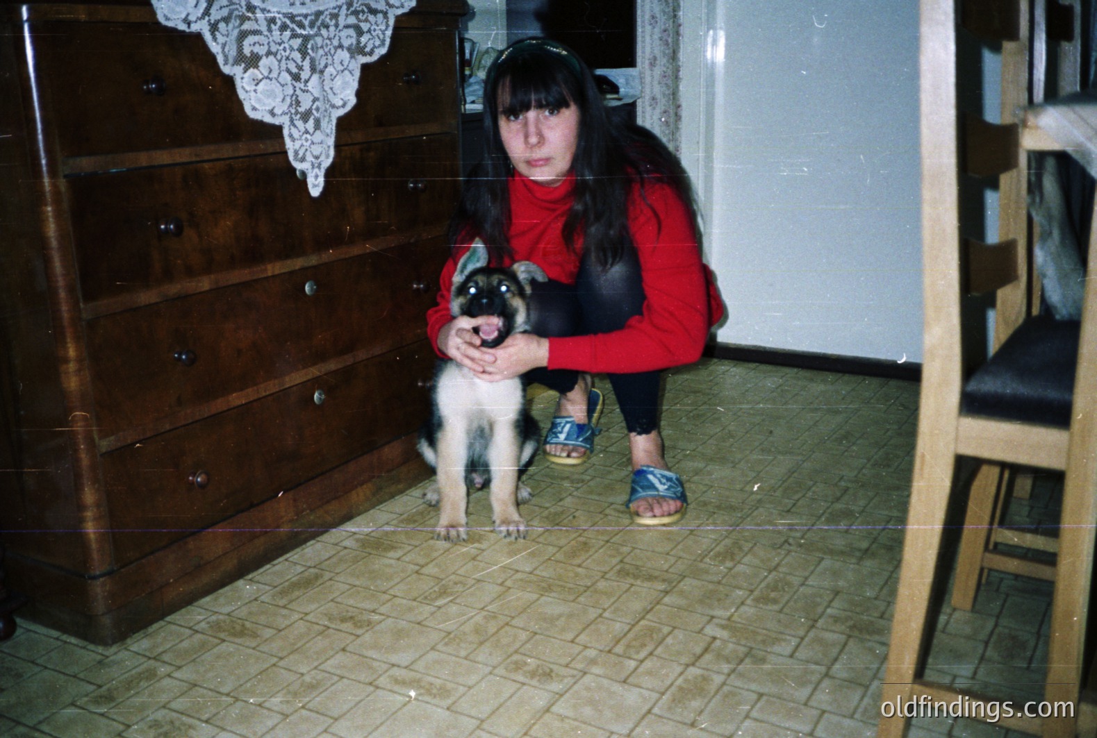 A young woman in a red turtleneck sweater and black leggings crouches, holding a small, black and white puppy. Interior setting with a patterned tile floor, dark wood furniture, and visible dated decor. Likely 1970s-80s style. The subject's style and the photographic quality suggest snapshot origin.