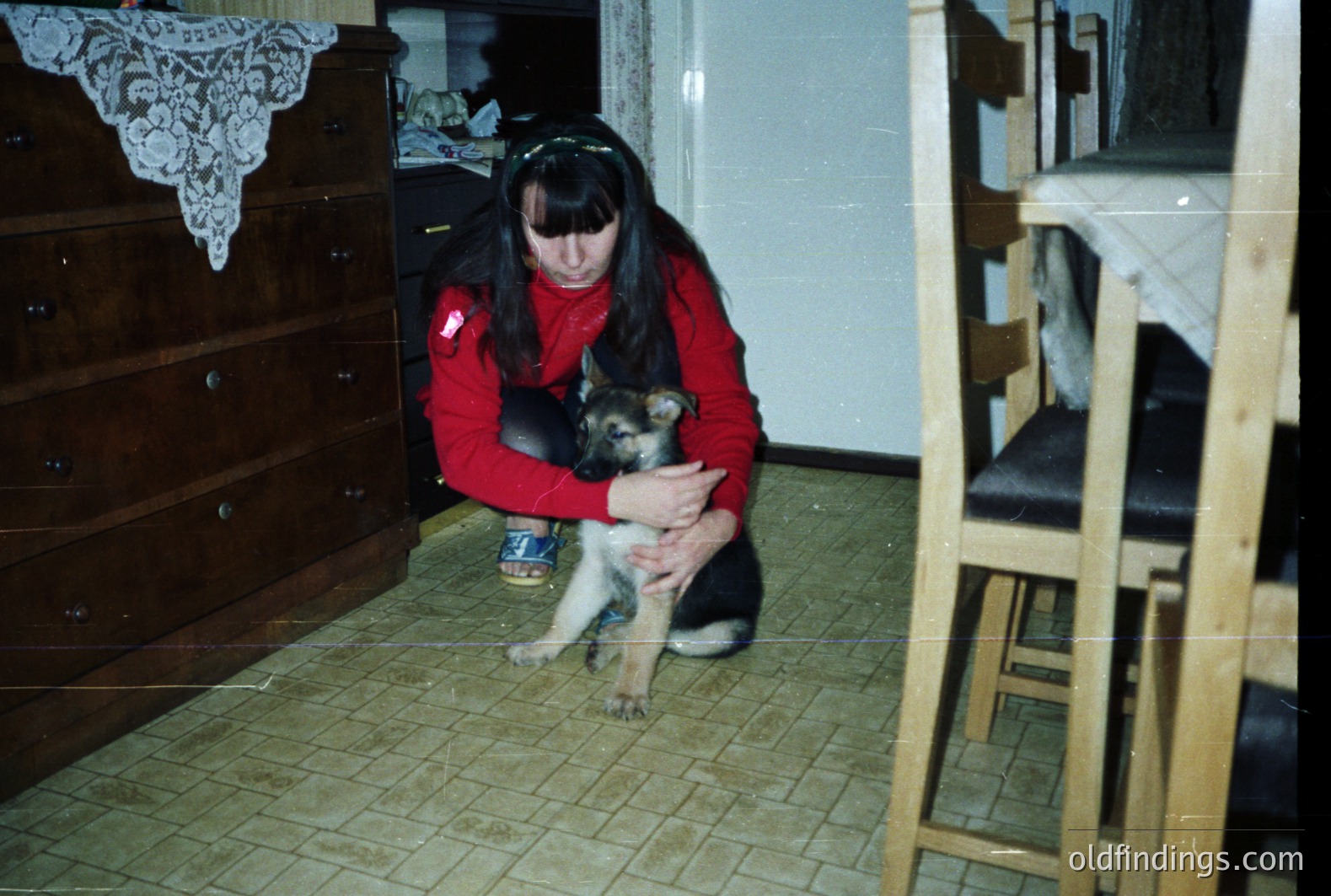 A young woman in a red sweater kneels, cradling a German Shepherd puppy indoors. Visible are a dresser with lace detailing, a stacked chair, and patterned linoleum flooring. Likely taken in the 1970s-1980s, based on the aesthetic. A can of food sits on the floor nearby.