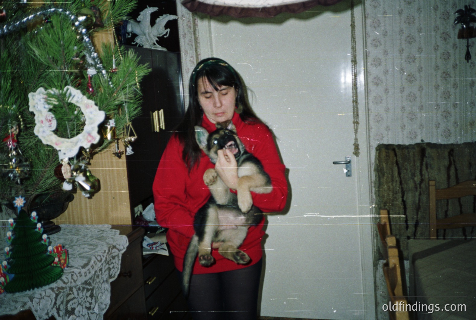 A young woman cradles a fluffy, long-haired kitten in her arms, posed near a decorated Christmas tree. The setting appears to be an older, possibly Eastern European home, evidenced by the patterned wallpaper & dark wood furniture. Likely taken in the 1980s, capturing a domestic, candid moment.