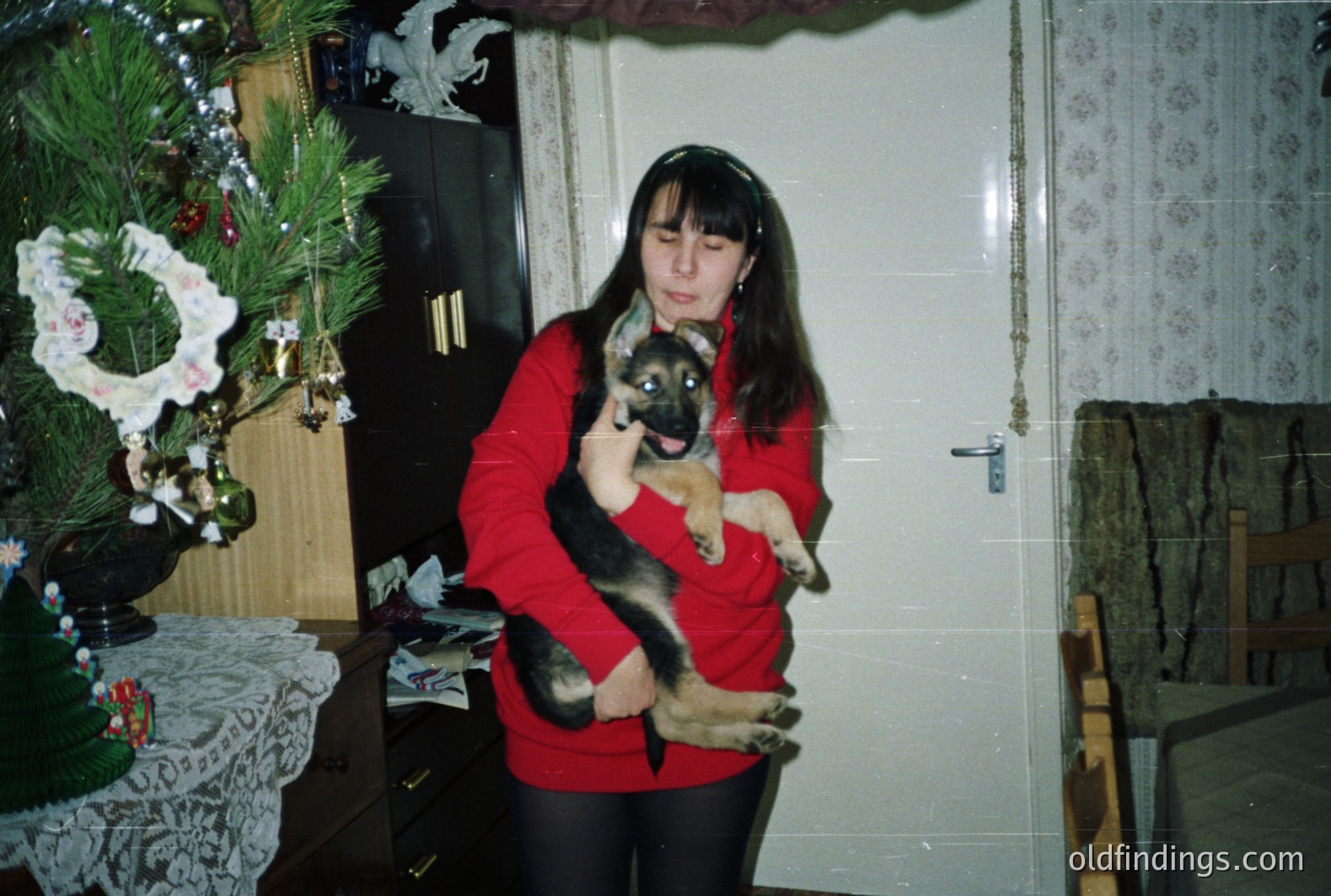 A young woman in a red sweater holds a German Shepherd puppy amidst a festive, likely Christmas, setting. Detailed lace tablecloth & decorated pine suggest a domestic interior. The grainy quality hints at a 1970s or 80s snapshot. Likely Eastern European, perhaps Bulgaria.