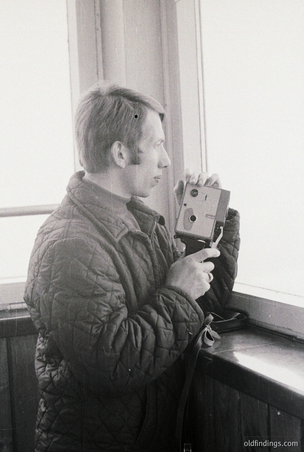 Man in a quilted jacket operates a vintage rangefinder camera, positioned near a window. Likely a candid shot, capturing a moment of photography in a domestic or travel setting. The style and dress suggest a 1960s or 1970s timeframe.