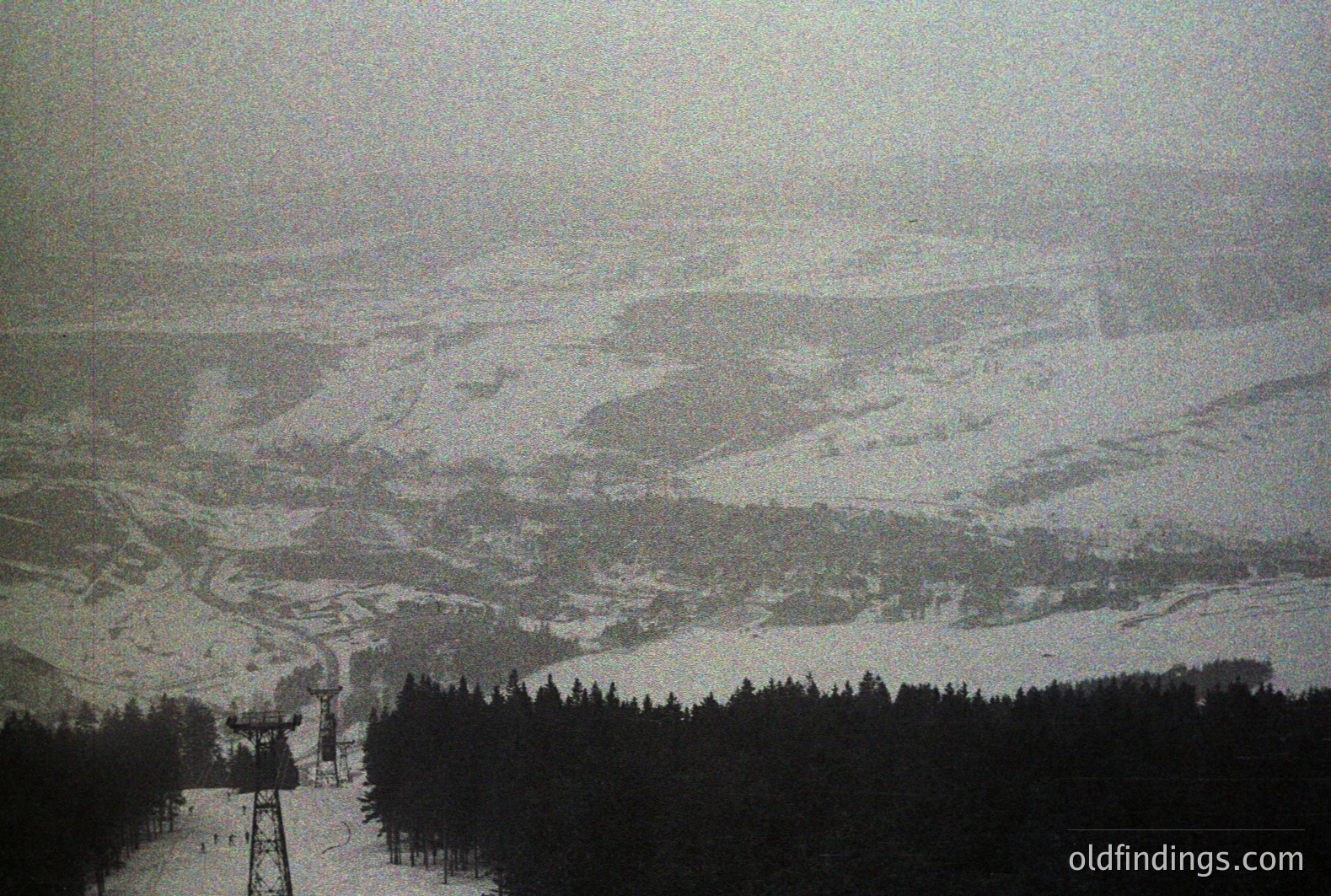 An aerial view captures a snow-covered alpine landscape featuring a ski lift tower and groomed slopes. Dense evergreen forest dominates the immediate foreground. Heavily textured, possibly aged, photographic print. Likely depicts a ski resort.