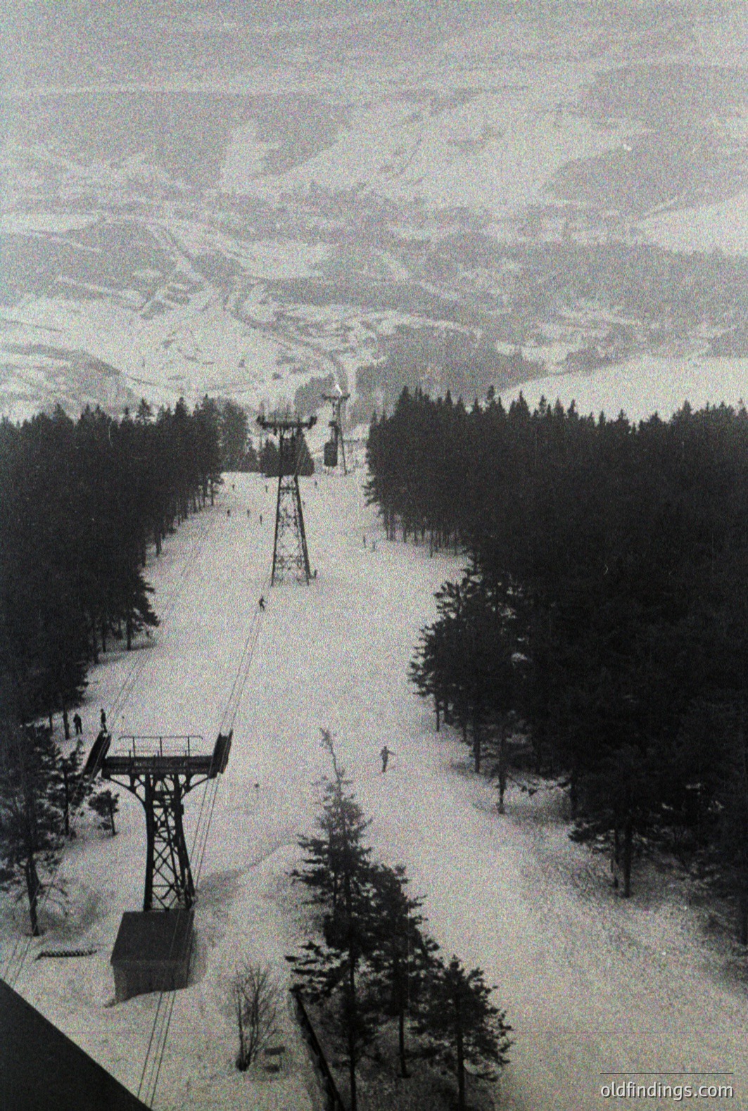 Striking view of a ski lift ascending a steep, snow-covered slope through dense pine forest. Distant mountain peaks are visible through the overcast sky. Likely a promotional image for a ski resort, potentially from the 1960s-1970s. A possible reference for design or historical research.