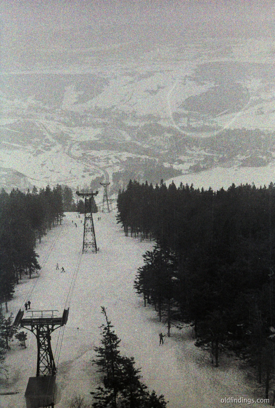 Monochrome view of a ski lift ascending a snow-covered slope. Evergreen trees line the lower portion, giving way to a wide, open ski run. Distant, snow-covered mountains fill the background. Likely a European location, mid-20th century. Potential archival value for ski tourism history.
