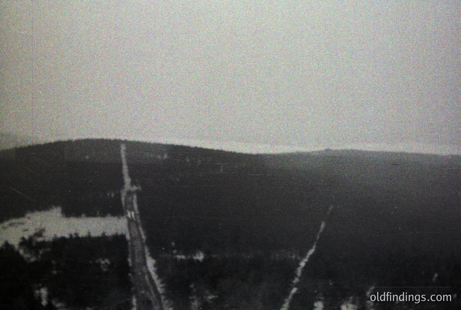 A high-angle view captures a ski lift ascending a forested, snow-covered slope. The stark monochrome highlights the infrastructure and landscape’s textures. Likely 1960s-70s, suggesting a historical alpine tourism development. The image's aesthetic offers design reference value.
