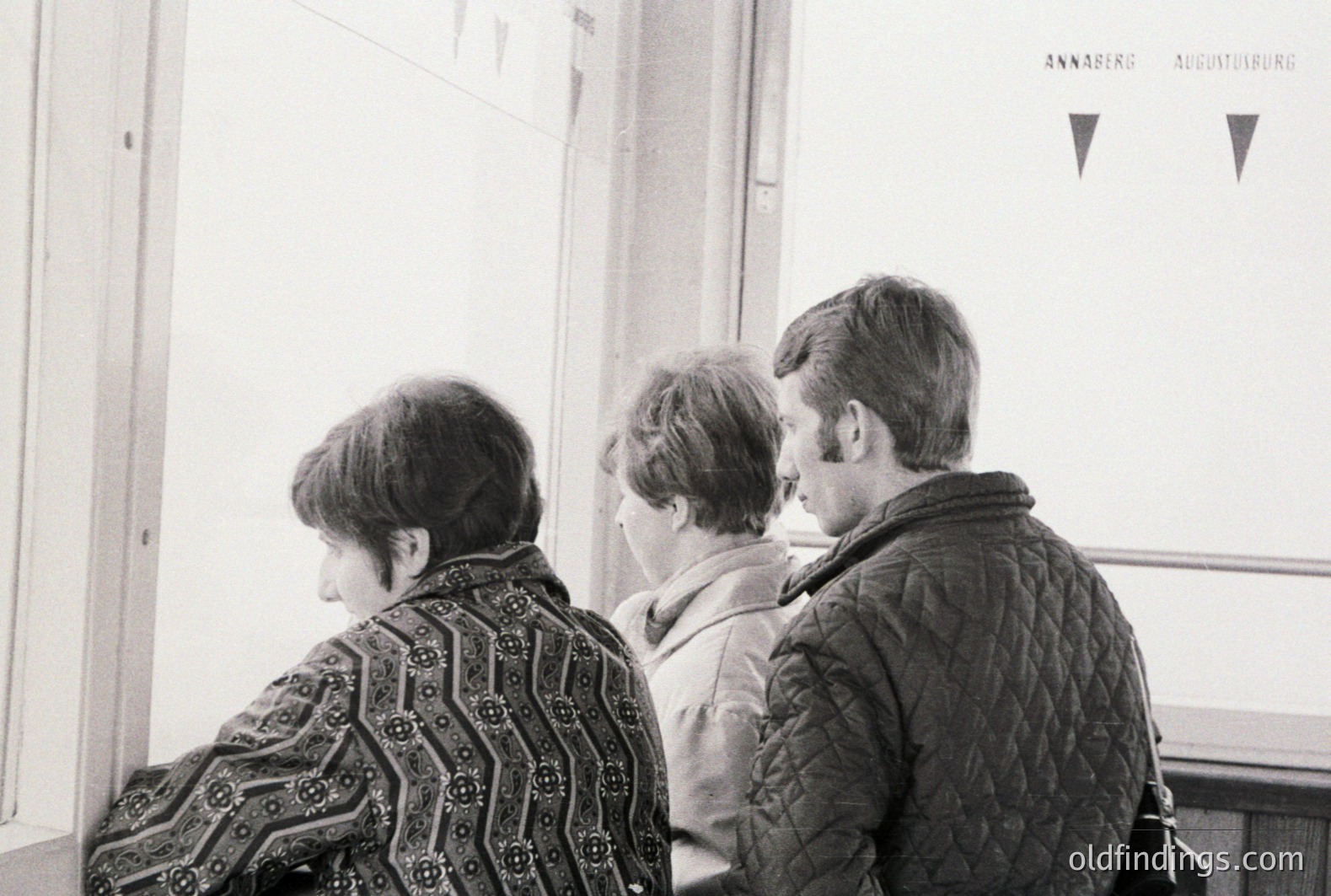 Three figures viewed from behind, likely on a public transportation vehicle; the older woman in patterned jacket, followed by a child and young man with shoulder bag. Minimalist signage indicating "Amberg" & "Augsburg" is visible. Likely 1970s style. A study in quiet observation.