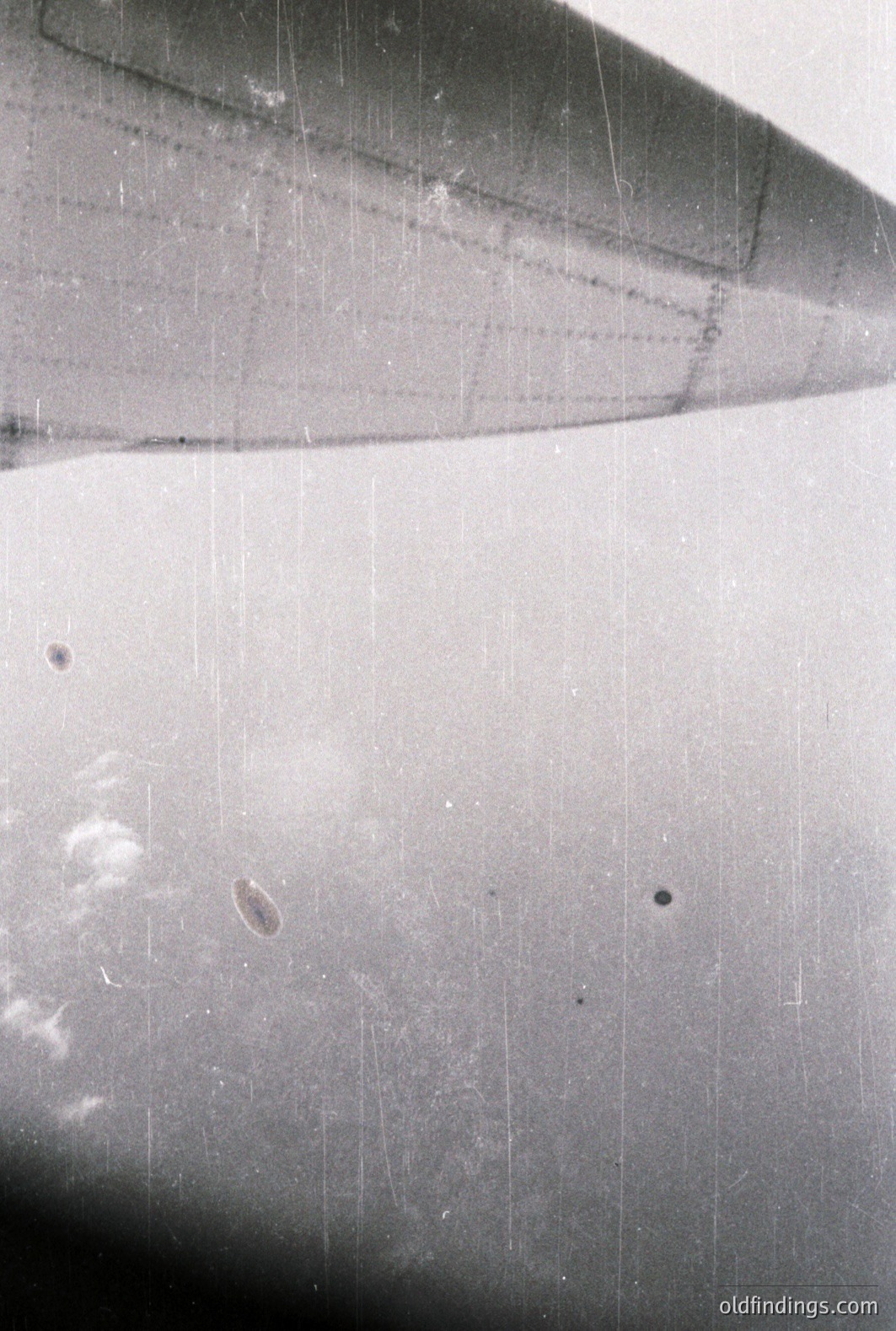 Close-up image showing the wing surface of an aircraft, likely a commercial jet. Visible weathering and streaking suggest prolonged exposure. Numerous scratches and minor imperfections across the metal finish. Possible rivets are visible. Reflective qualities indicate a bright, sunny day. Likely mid-20th century.