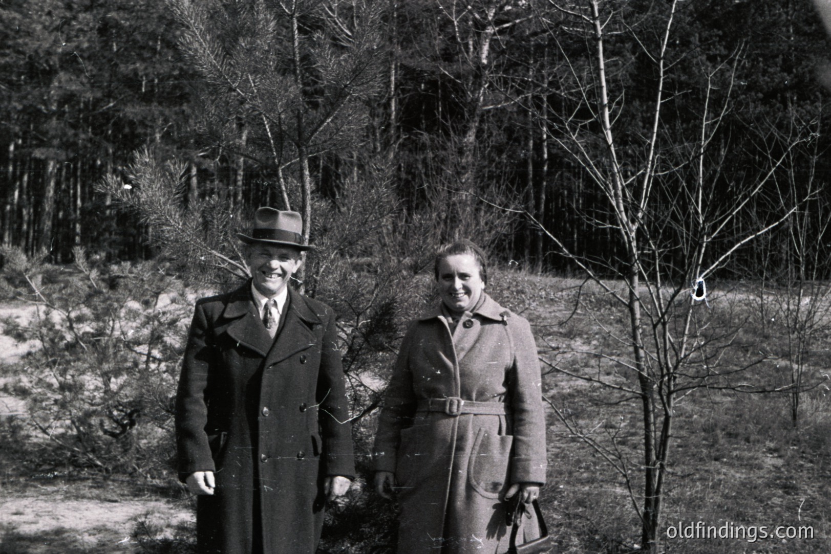 A formally-dressed man and woman stand outdoors, likely in a park or forest setting. Both wear long overcoats and hats, suggesting a colder climate or era. The man holds a tie, while the woman carries a dark satchel. Trees are visible in the background.